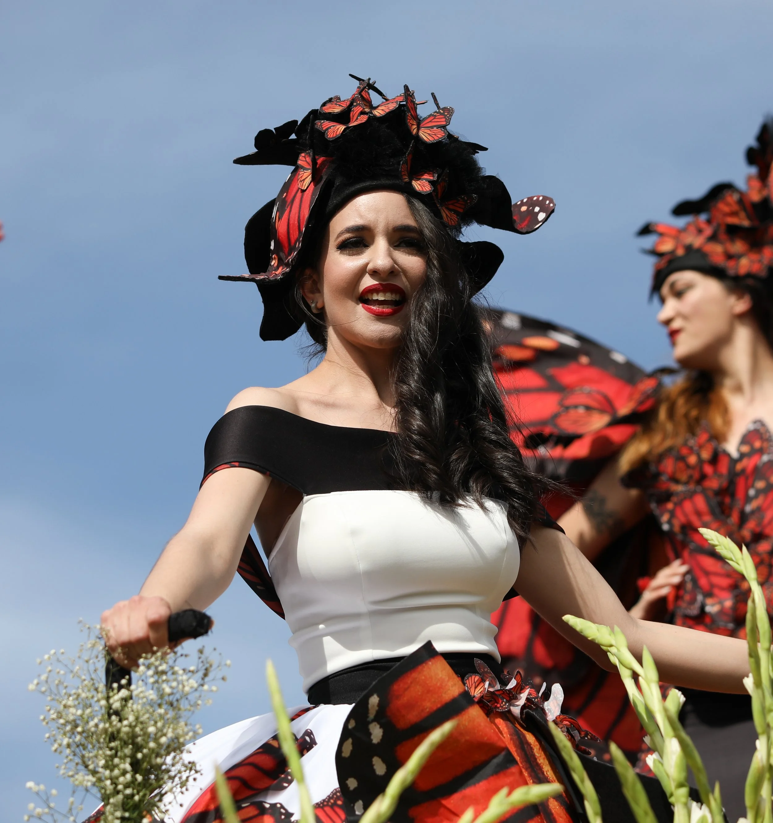 Women participating in a cultural or festive event, wearing elaborate black and red butterfly-themed costumes and hats, in an outdoor setting under a clear sky.