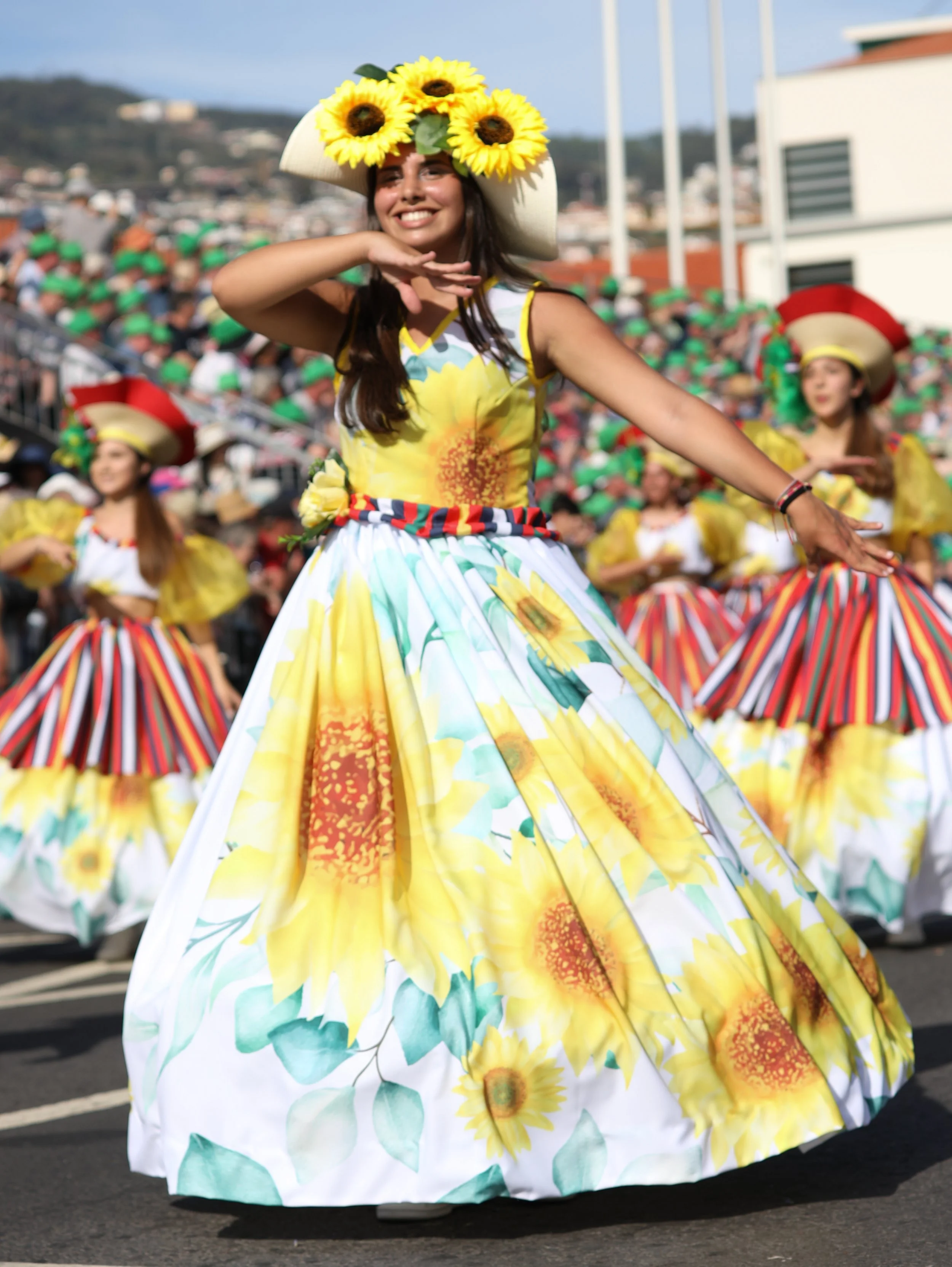 Young woman smiling and dancing in a colorful sunflower-themed dress and large sunflower hat during a parade; other dancers in similar outfits are in the background.