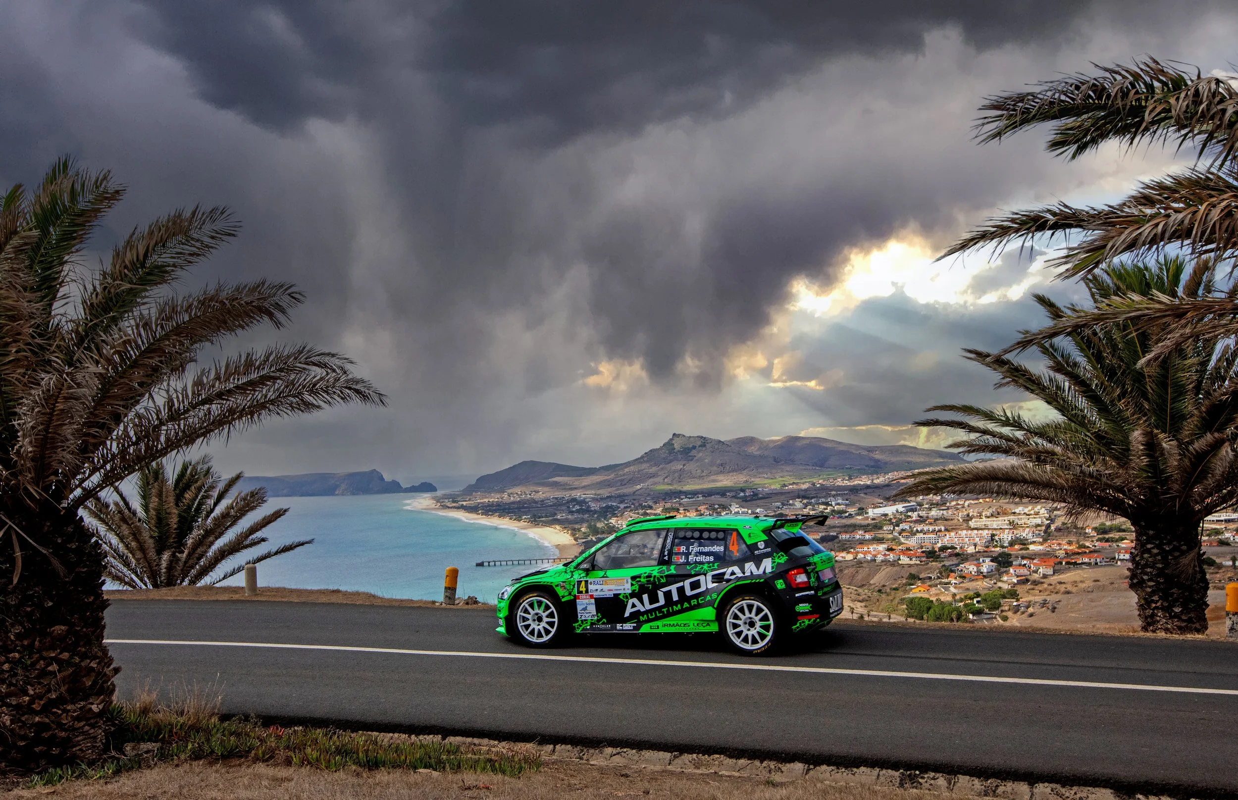 A green and black rally car driving on a coastal road with palm trees on both sides and a dramatic cloudy sky overhead, overlooking the ocean and a town below.