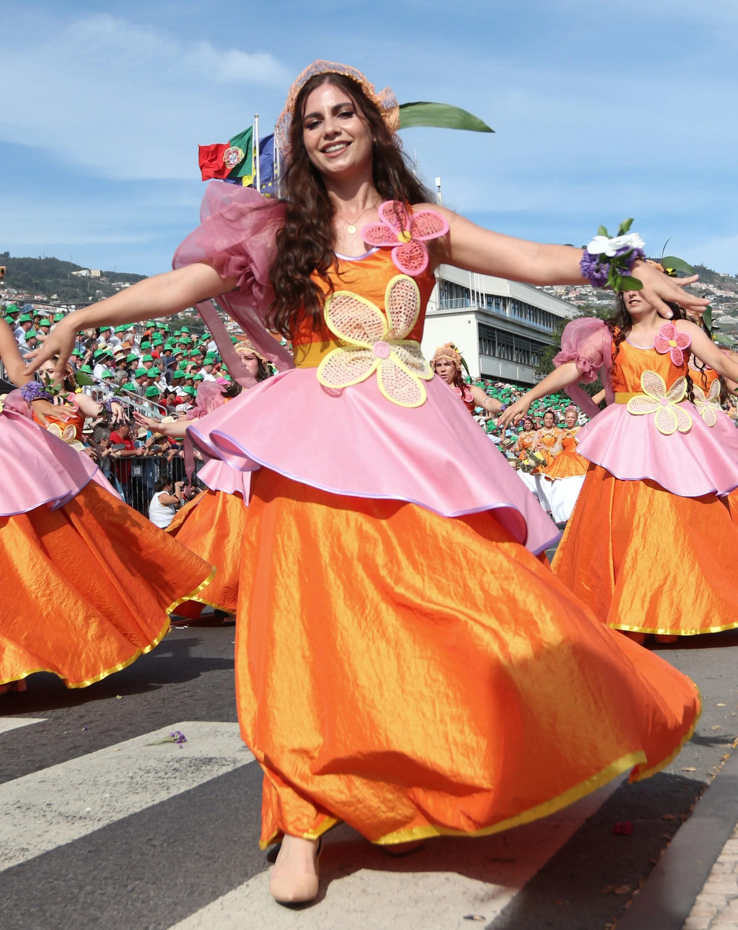 Women dancing in colorful traditional costumes during a parade, with a crowd of spectators and flags in the background.