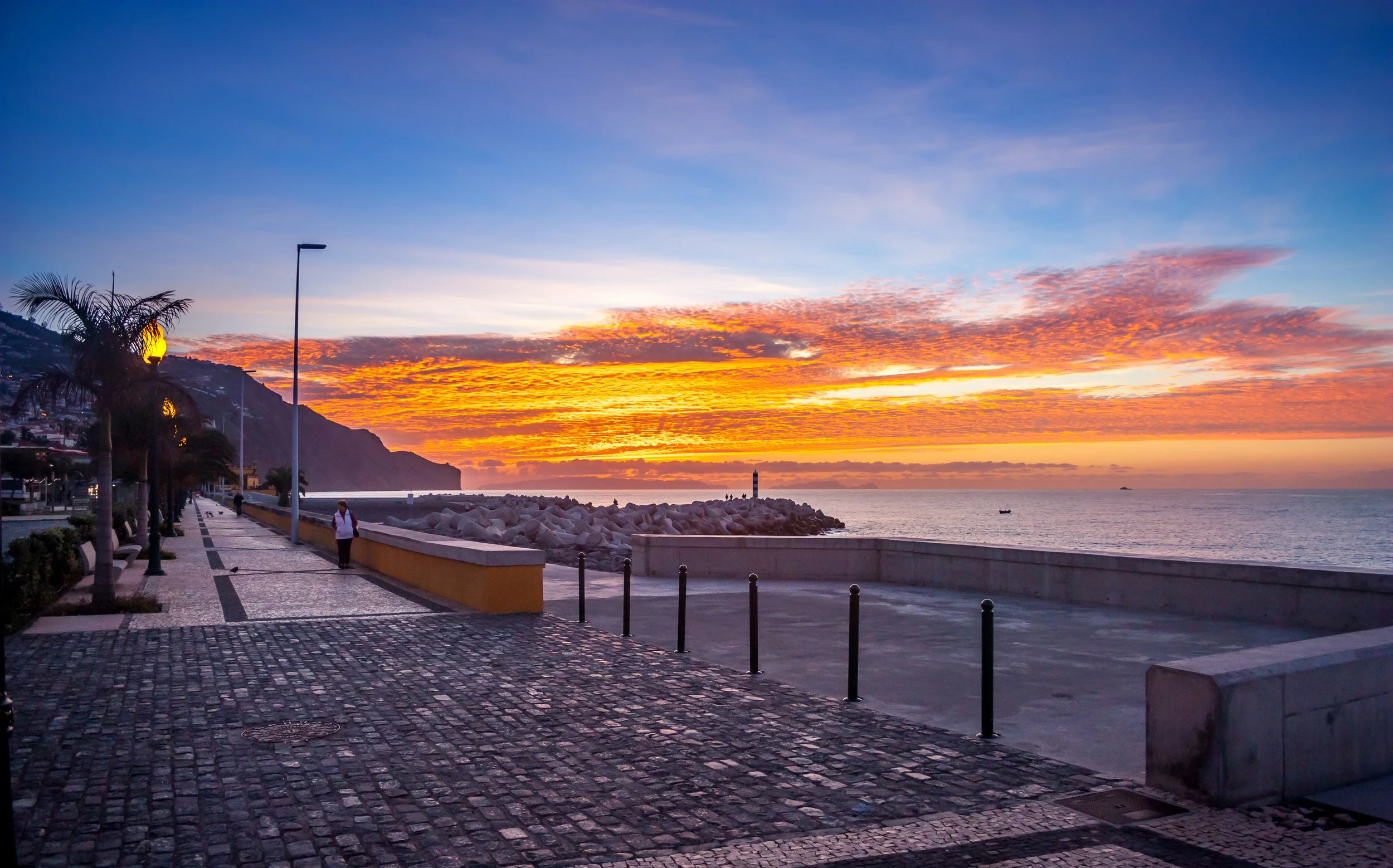Sunset over a seaside promenade with a cobblestone walkway, palm trees, street lamps, and a breakwater with rocks and a lighthouse in the distance.