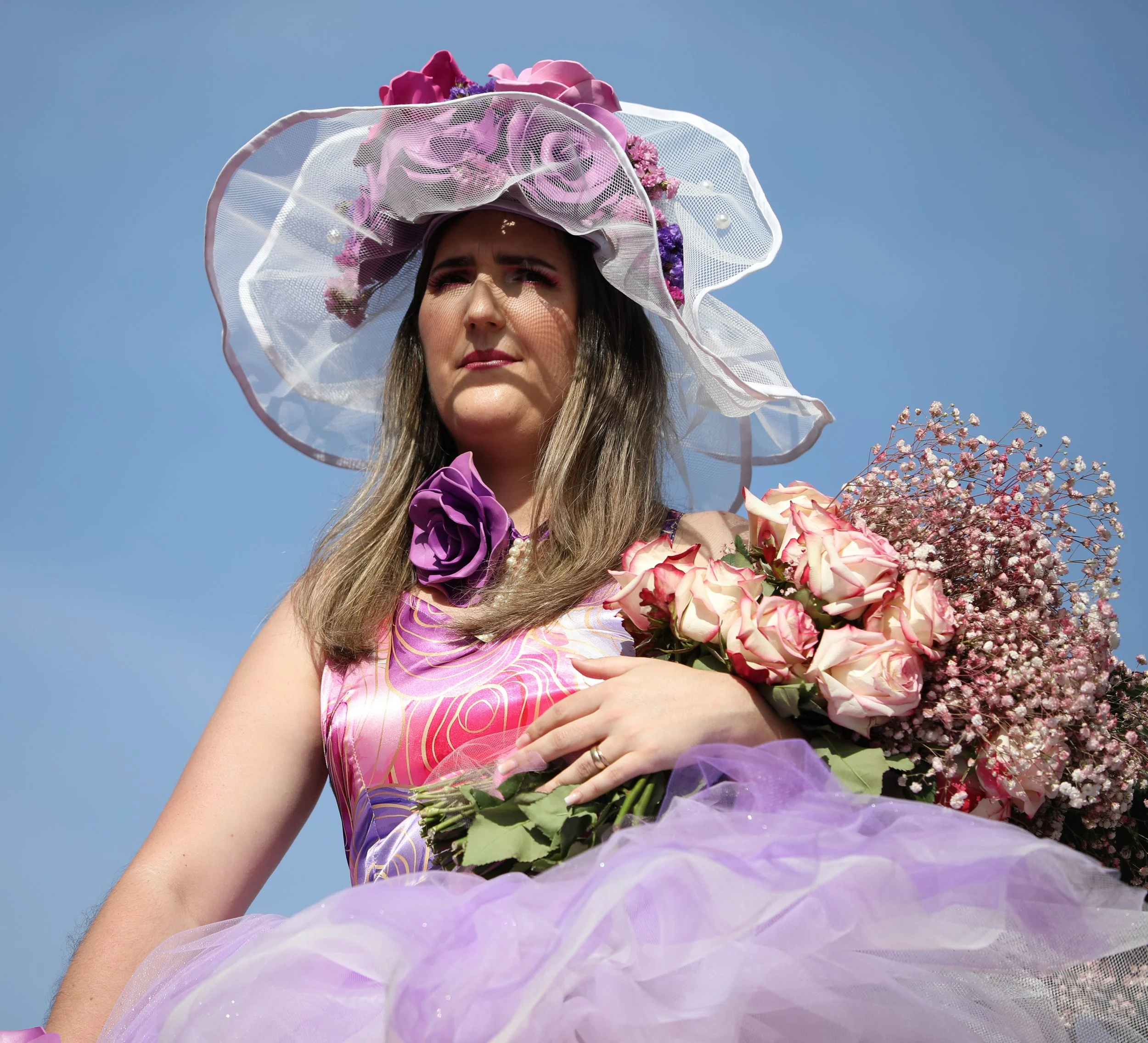 A woman wearing a large, floral, pink and purple hat with a veil, holding a bouquet of pink roses and baby's breath, against a bright blue sky.
