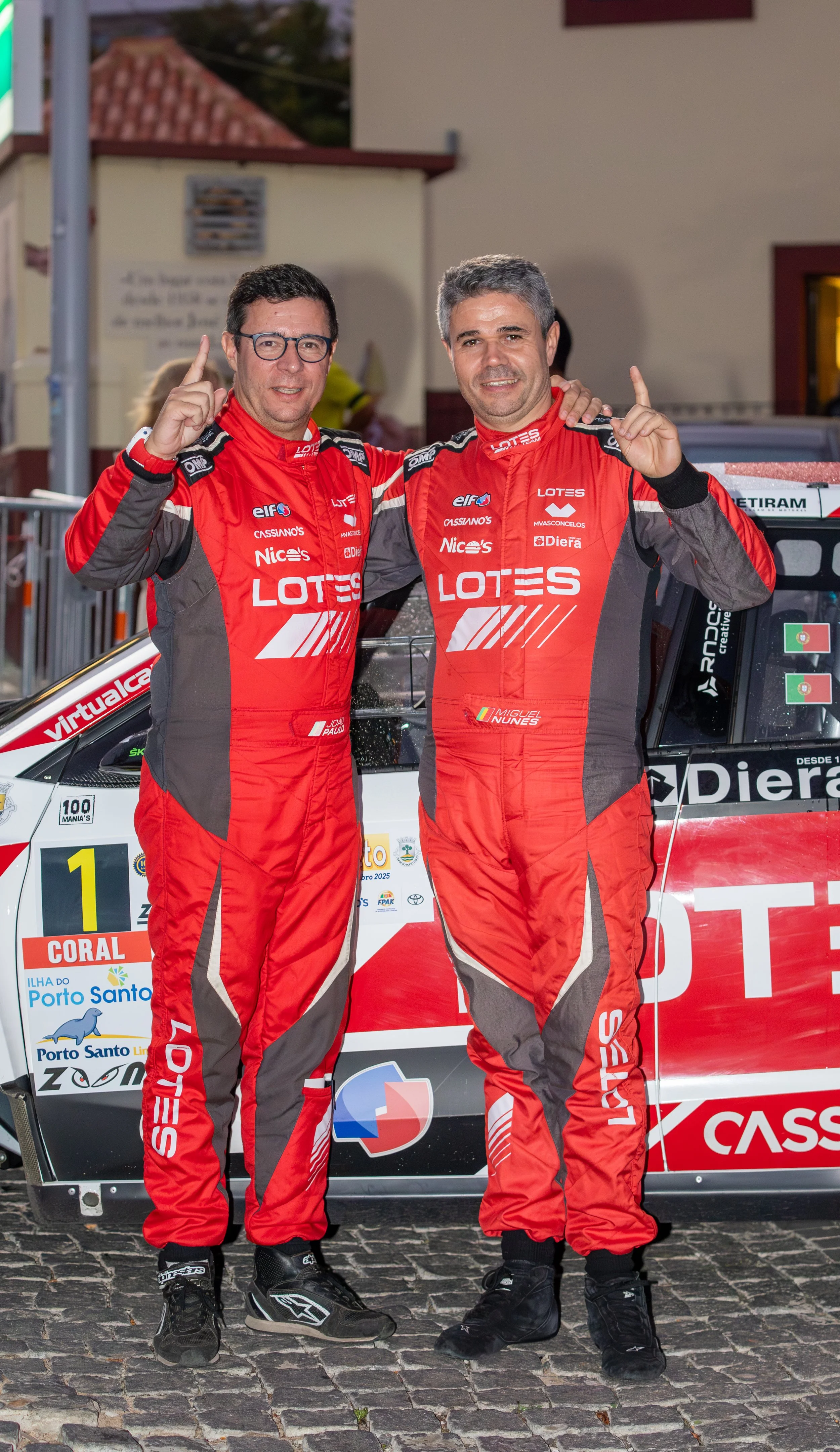Two race car drivers in red and gray racing suits standing in front of a rally car, celebrating and holding up their index fingers.