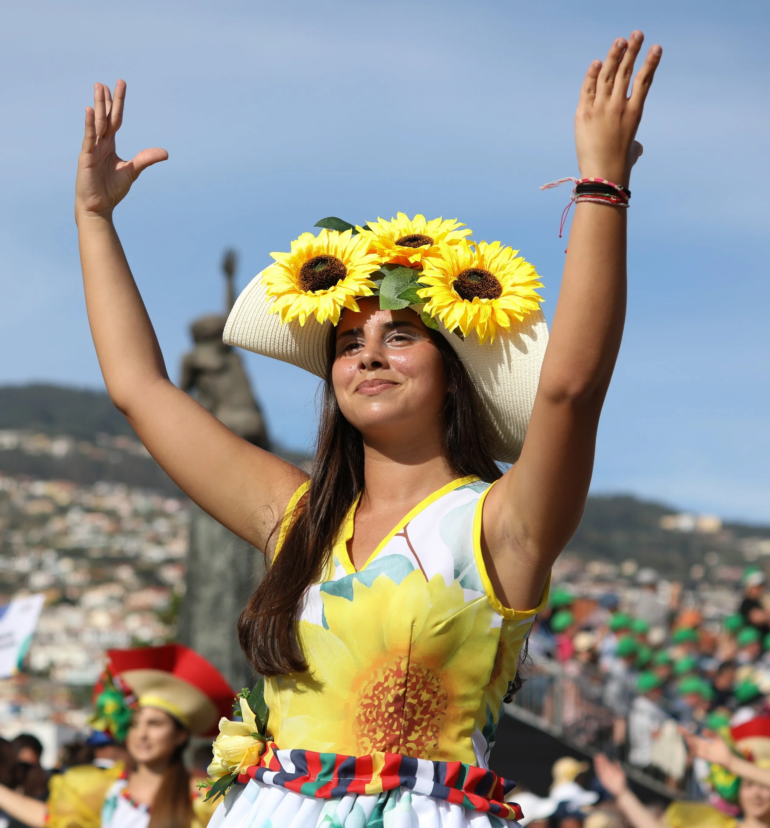 A woman wearing a large sunflower hat and a sunflower-themed dress participating in a cultural celebration or parade. She has her arms raised and appears to be enjoying the event, with a crowd and a hilly landscape in the background.