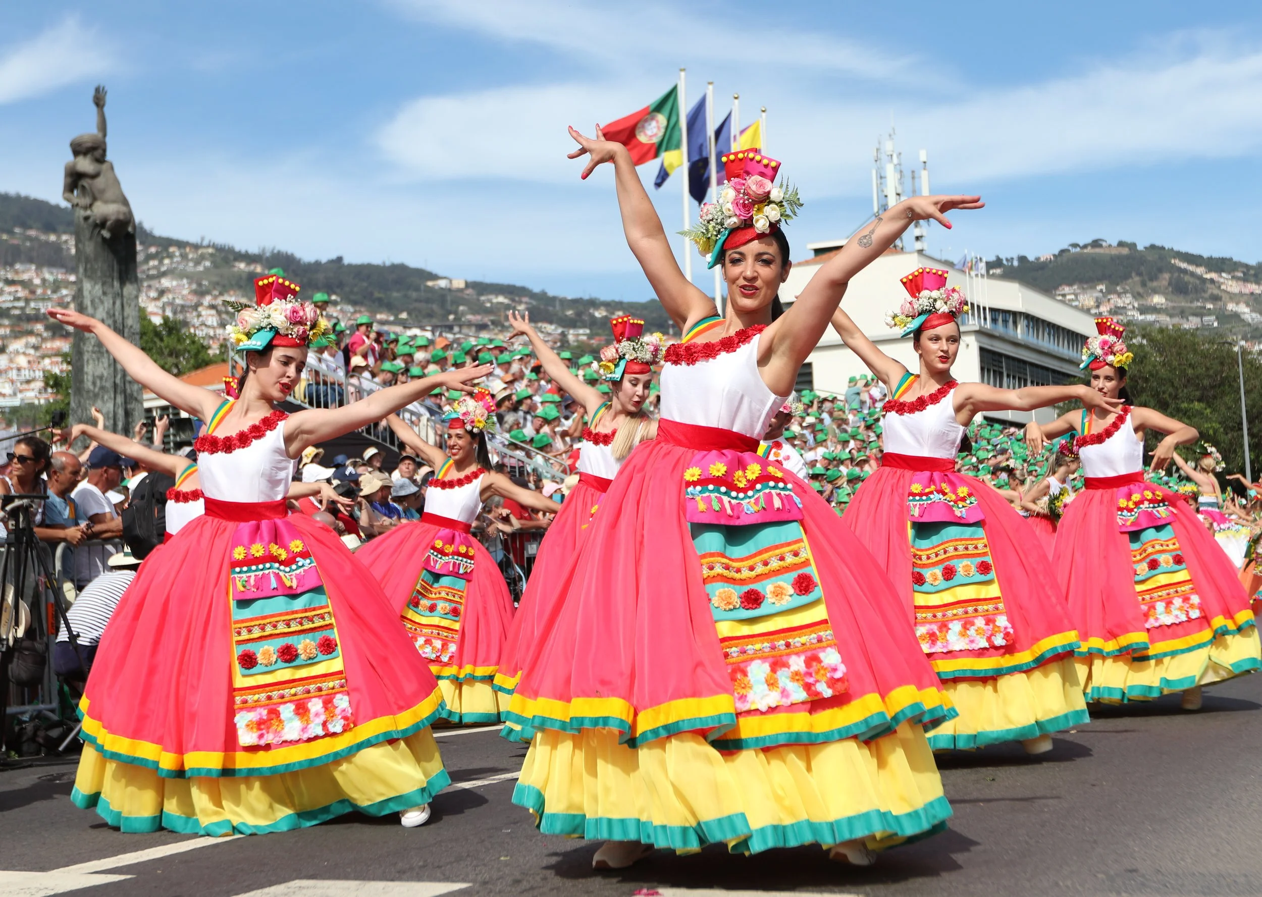 A group of women dressed in traditional colorful Mexican dance costumes performing in a parade with a crowd and flags in the background.