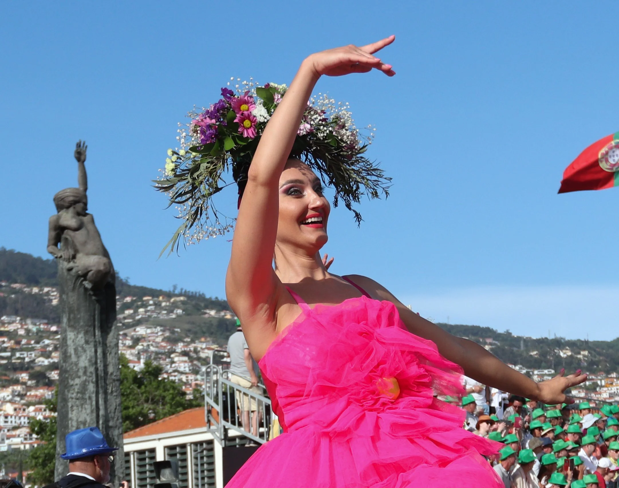 Woman dressed in a bright pink dress with a floral headpiece, dancing with arms raised outdoors during a festival or parade.