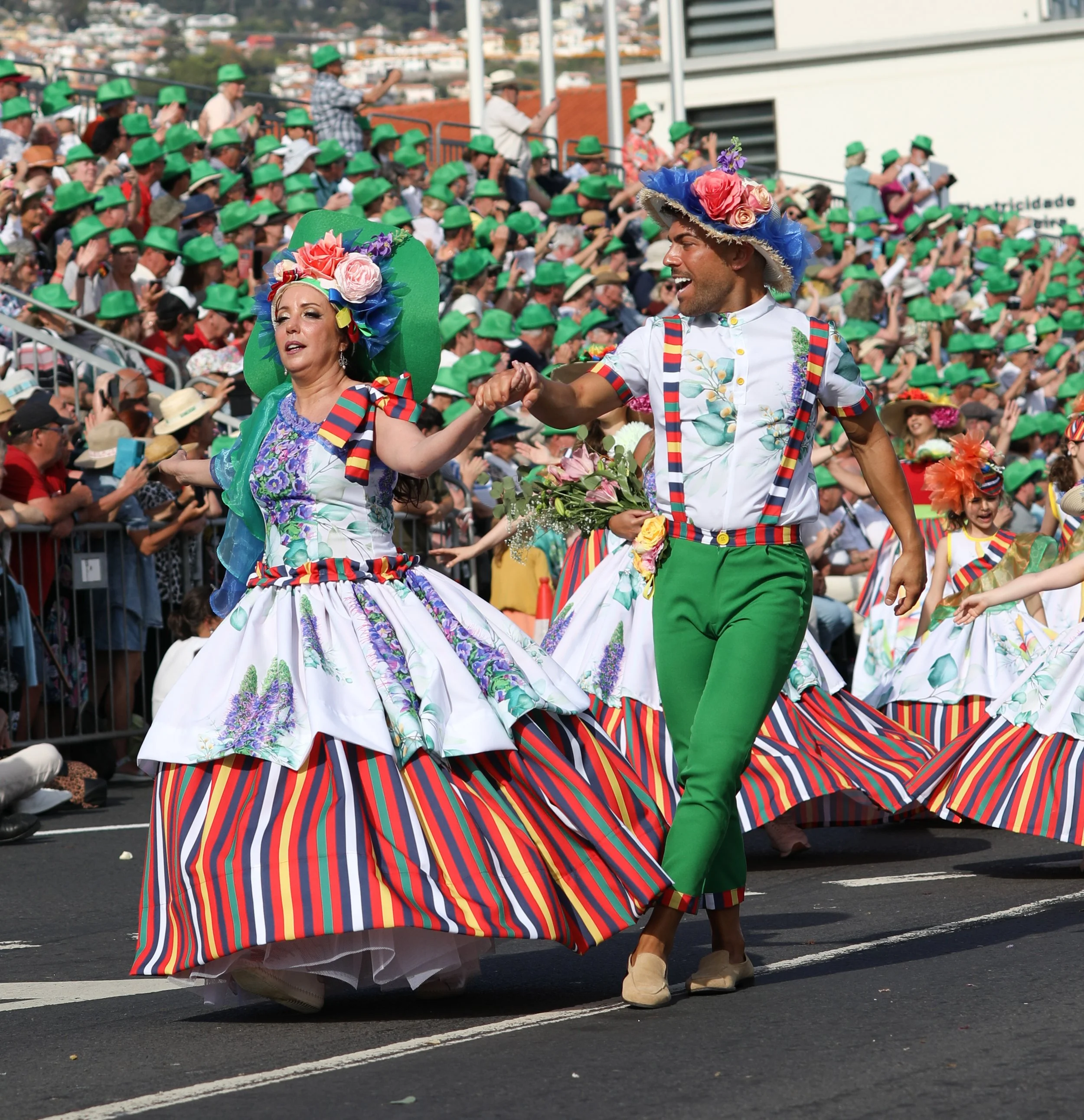 Couple dancing in colorful traditional costumes during a parade, with a large crowd of spectators wearing green hats in the background.