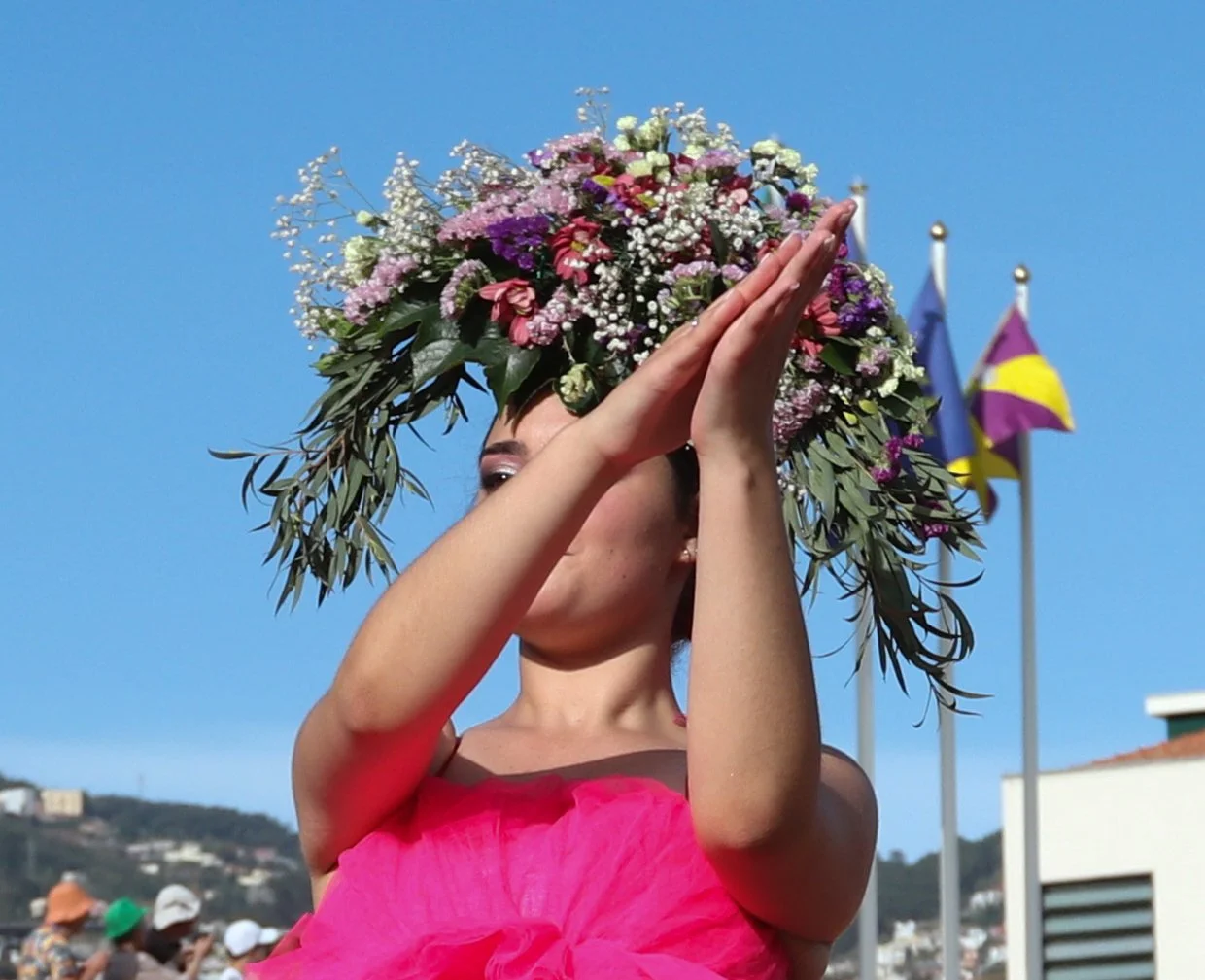 A woman wearing a pink dress and an elaborate floral headdress with various flowers and greenery, with flags in the background on a clear blue sky day.