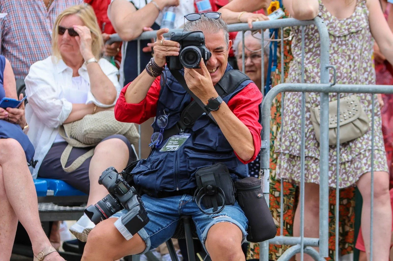 A man is sitting on a chair, taking a photograph with a camera. He has gray hair, is smiling, and is wearing a navy vest with a red sleeve shirt underneath. He has another camera hanging from his waist and is surrounded by a crowd of people at an outdoor event.