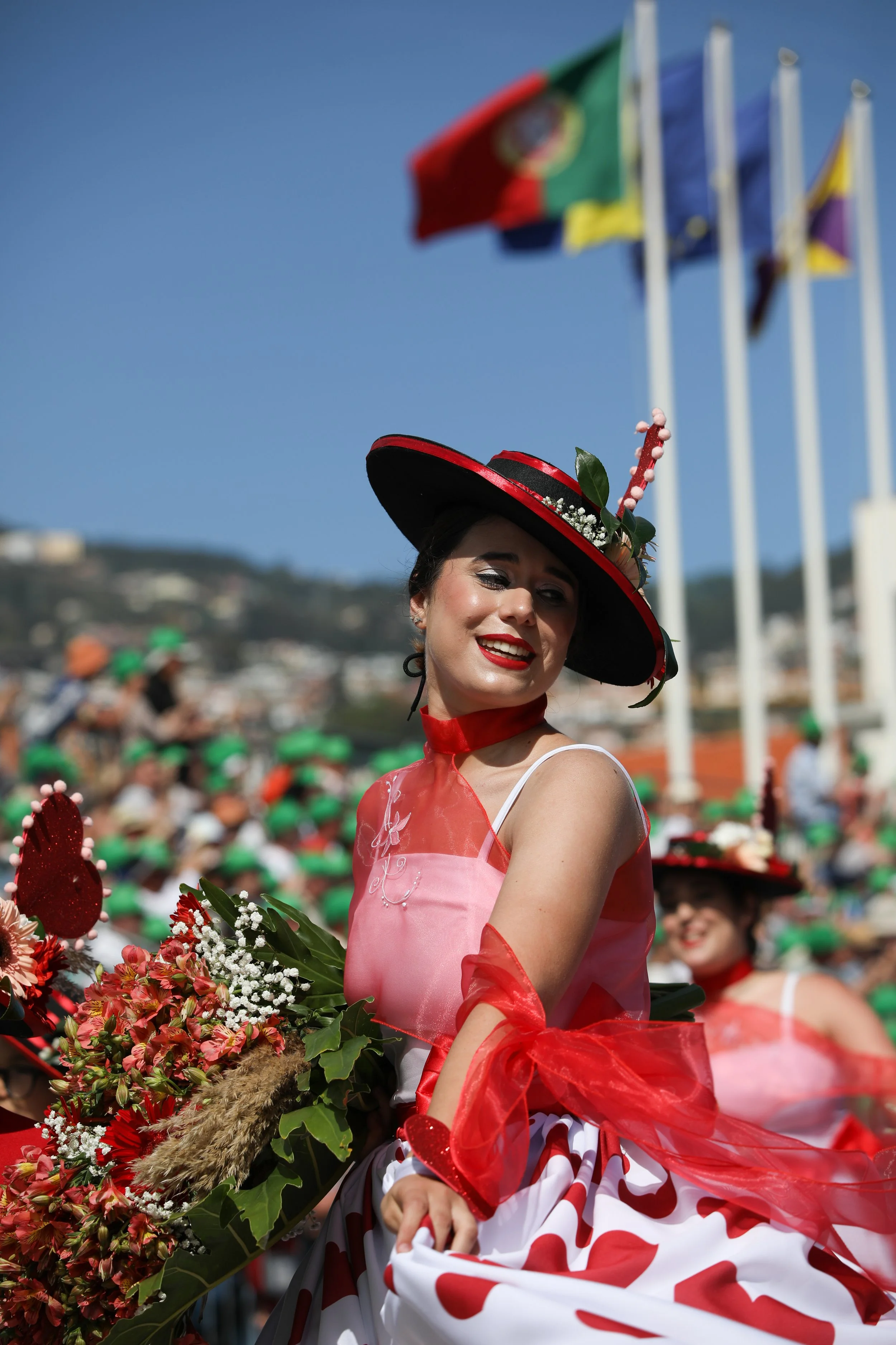 Woman in traditional dress holding a bouquet, smiling at a parade with flags in background.