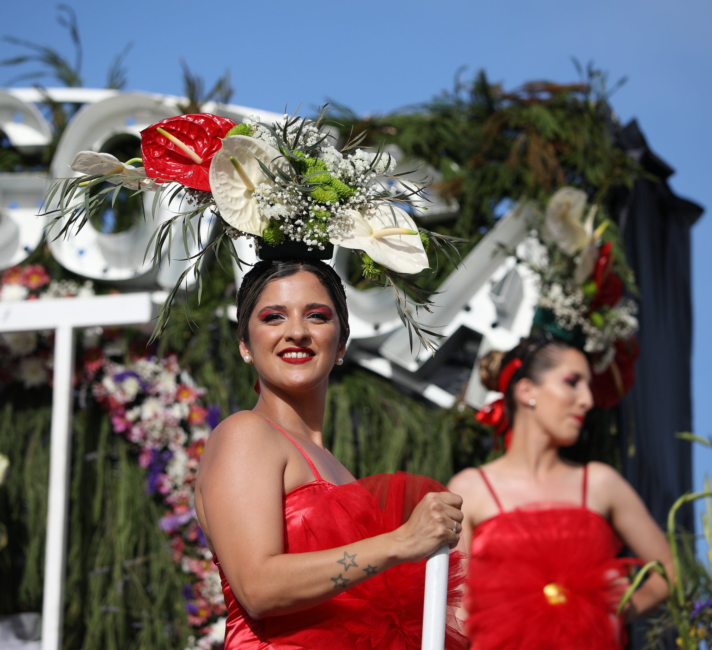 Two women dressed in red with elaborate floral headpieces on a parade float, with a blue sky in the background.