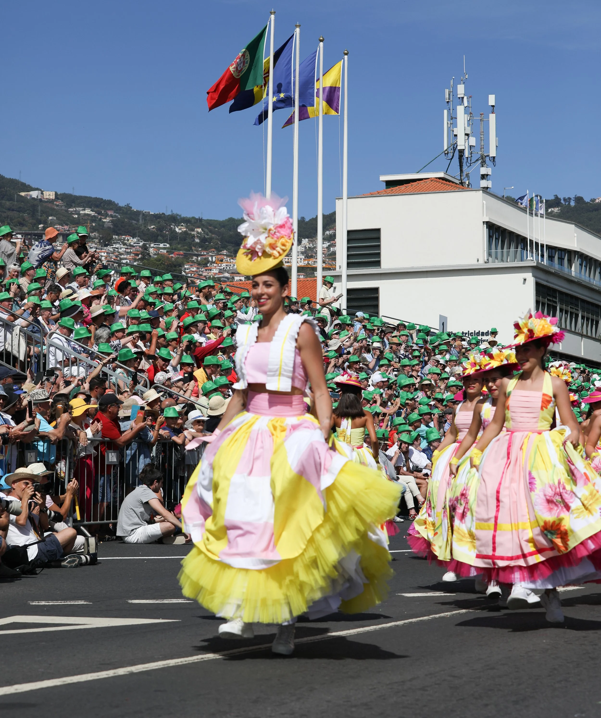 Colorful parade in Portugal with women in traditional dresses with large floral hats, spectators in hats and sunglasses, and flags of Portugal, European Union, and regional flags in the background.