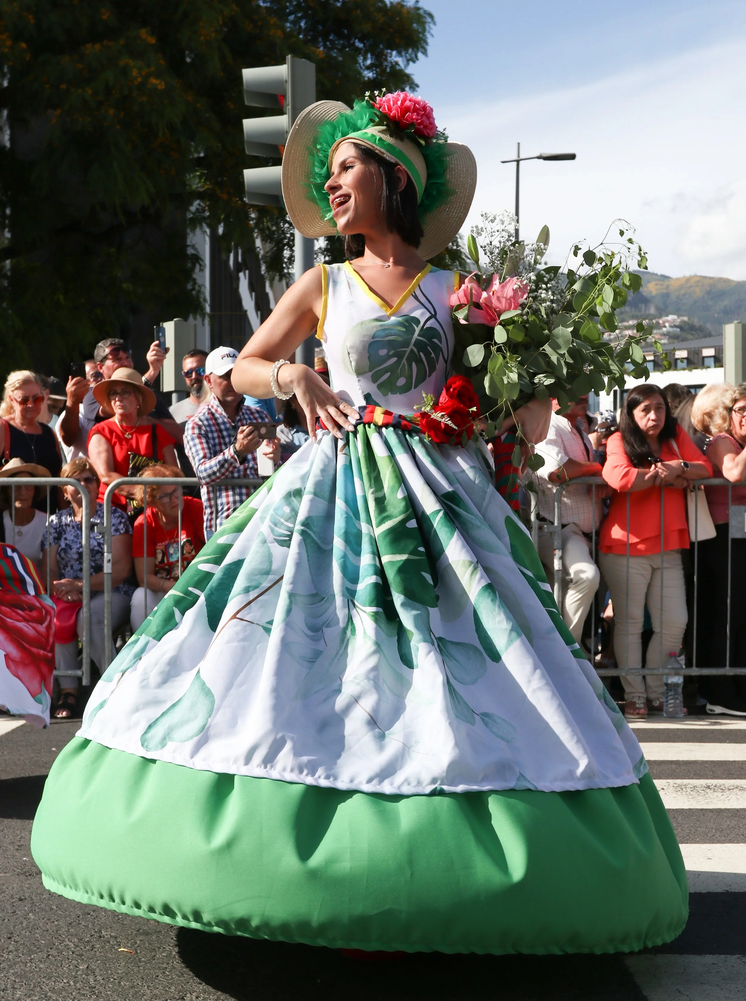 A woman dressed in a vibrant, oversized floral-printed dress with a matching wide-brimmed hat decorated with a pink flower, during a parade, holding a bouquet of flowers, with onlookers behind her on a city street.