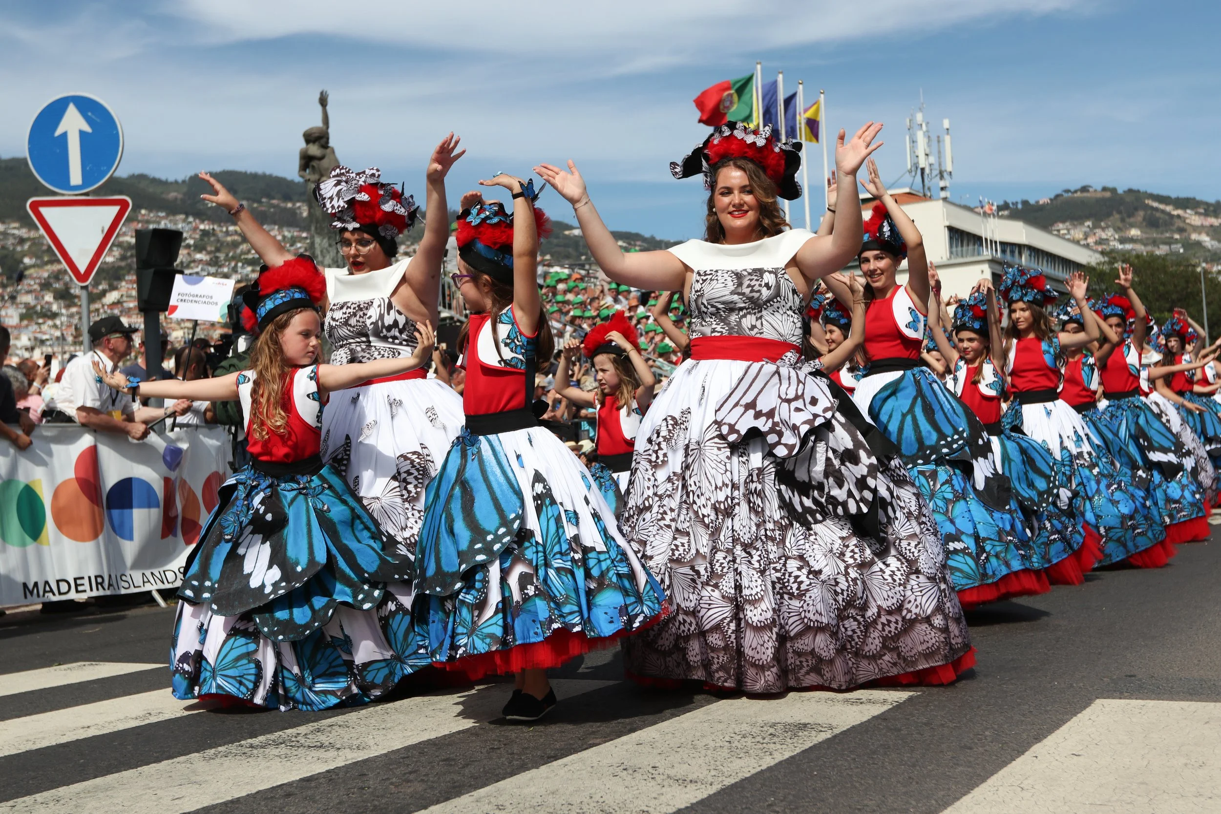 Group of women and children dressed in traditional butterfly-themed costumes participating in a public parade on a city street, with flags and spectators in the background.