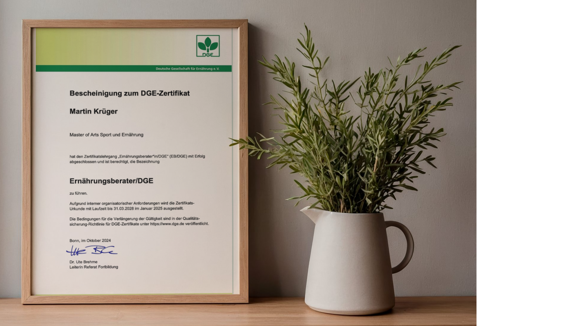 Zertifikat der Deutschen Gesellschaft für Ernährung (DGE) im Rahmen eines Rahmen, neben einer weißen Vase mit grünem Pflanzen. Das Zertifikat ist in einem Hol frame, auf einem Holztisch. Eine graue Wand im Hintergrund.