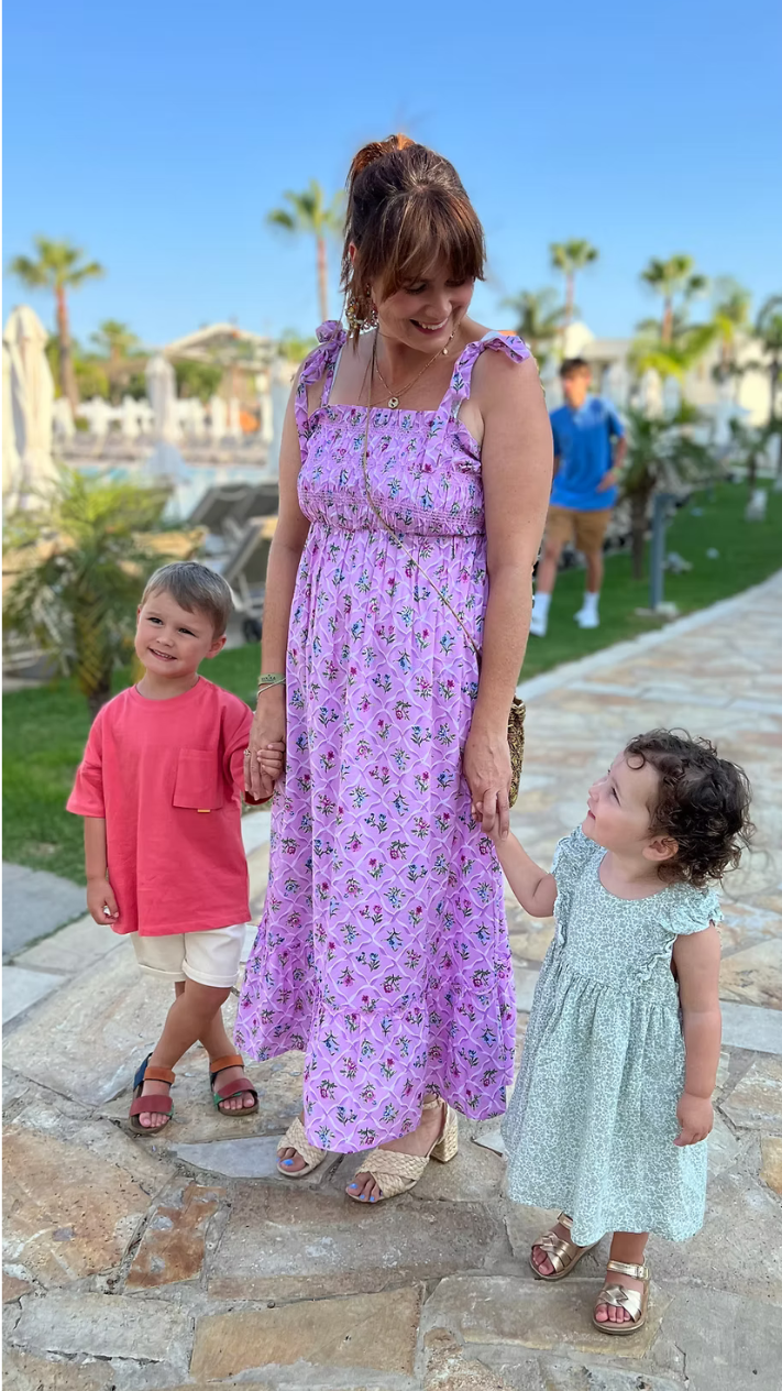 A woman in a purple floral dress holding hands with two children, a boy in a pink shirt and white shorts and a girl in a green dress, outside on a stone pathway with palm trees and blue sky in the background.