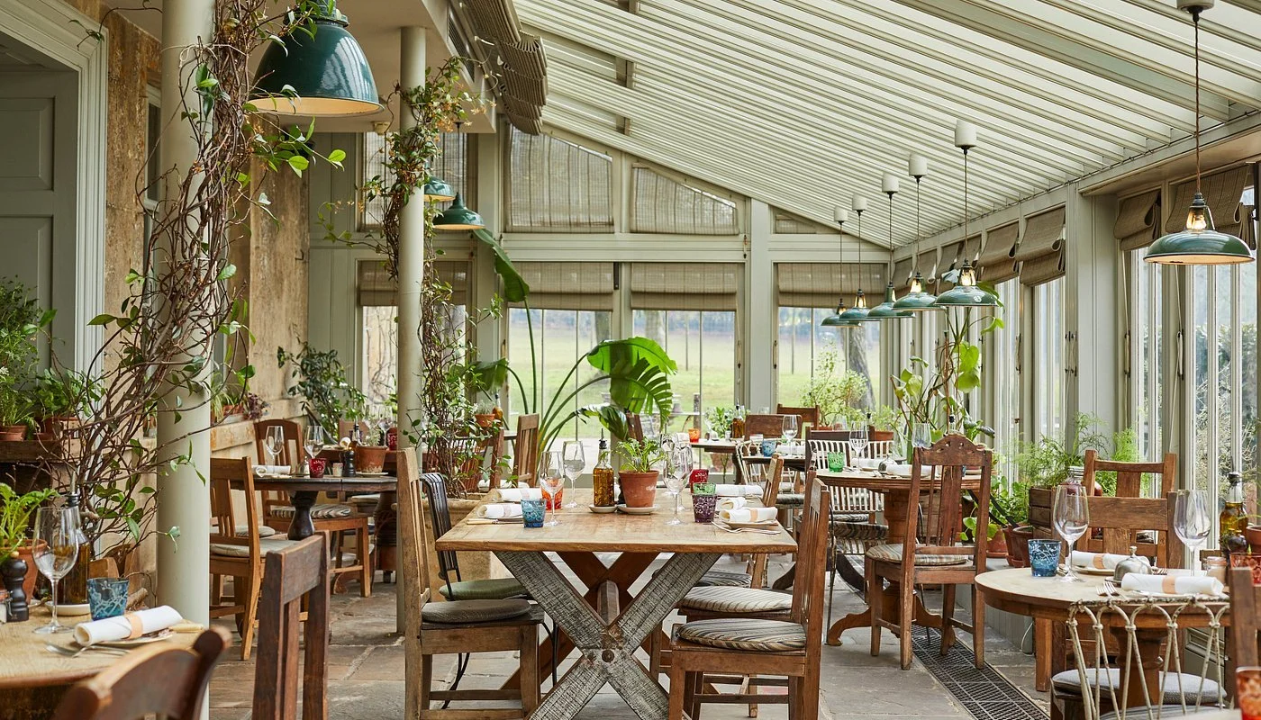 A sunroom with wooden tables and chairs, decorated with potted plants, and set with wine glasses, napkins, and colorful glassware, with large windows and a sloped ceiling.