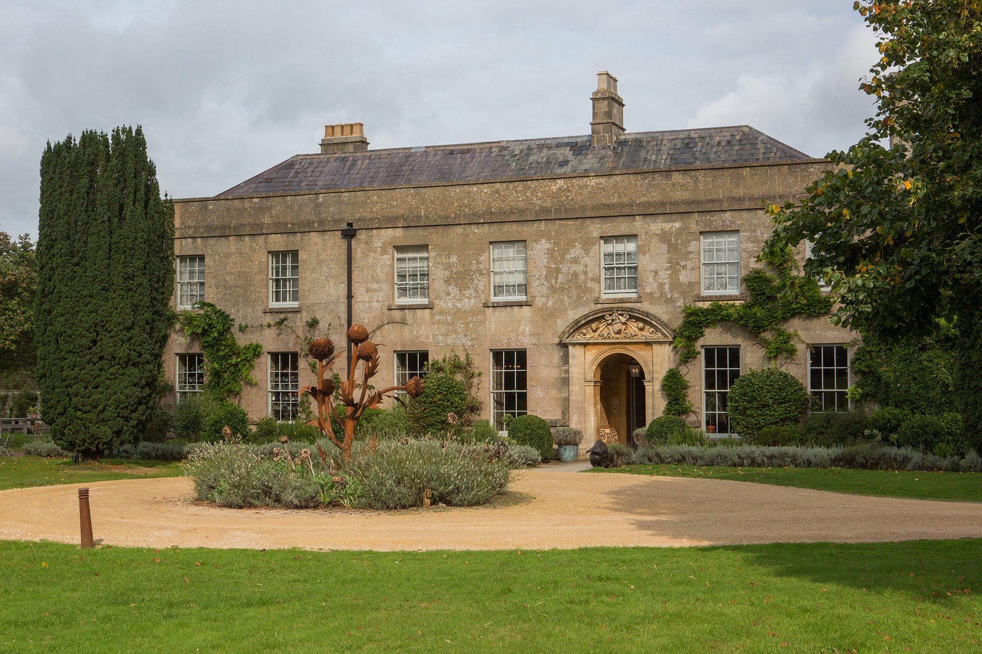 A large historic stone house with multiple windows and a decorative arched door surrounded by a landscaped garden with shrubs, trees, and a paved walkway.