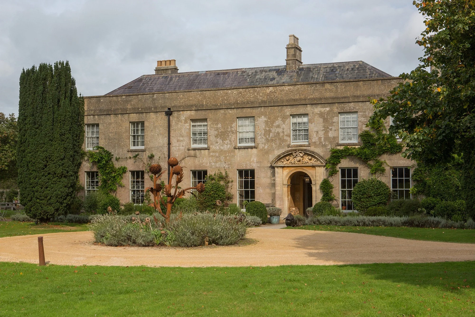 A large stone house with multiple windows, surrounded by greenery and a garden, under a cloudy sky.