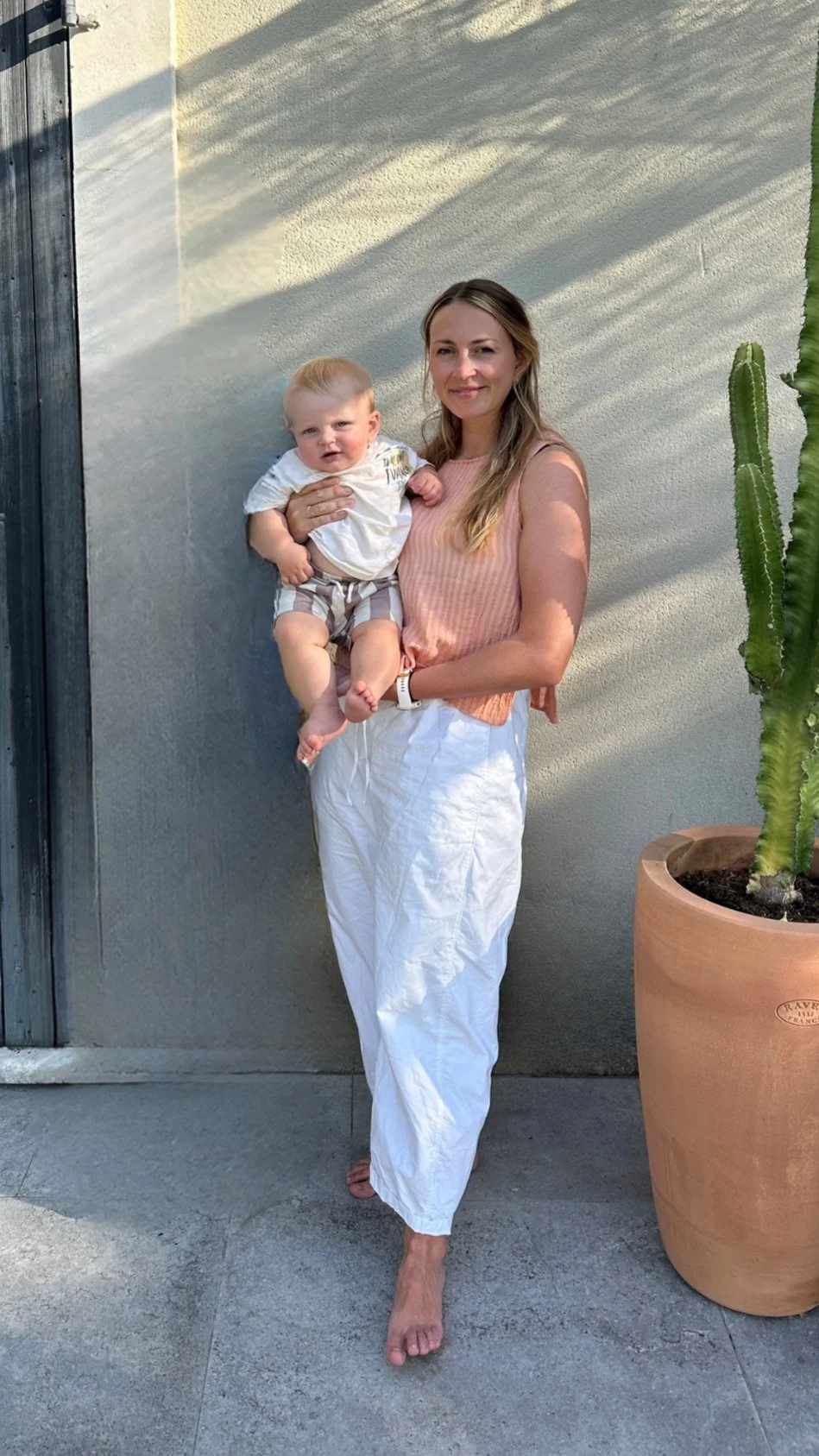 A woman holding a young child outdoors near a beige textured wall and a large potted cactus. The woman is smiling, wearing a peach-colored sleeveless top and white pants, in sunlight with shadows on the wall.
