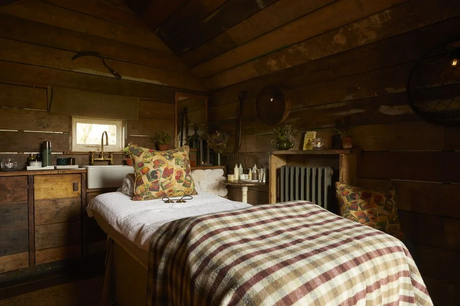 Cozy bedroom with wooden walls, a bed with a plaid blanket and colorful pillows, small window, and rustic decor.