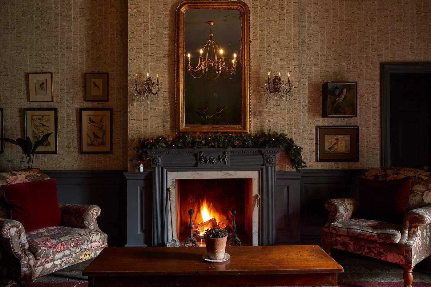 A cozy living room with a lit fireplace, decorated with greenery and candles, and framed bird artwork on the wallpapered walls. Two upholstered armchairs sit on either side of a wooden table in front of the fireplace.