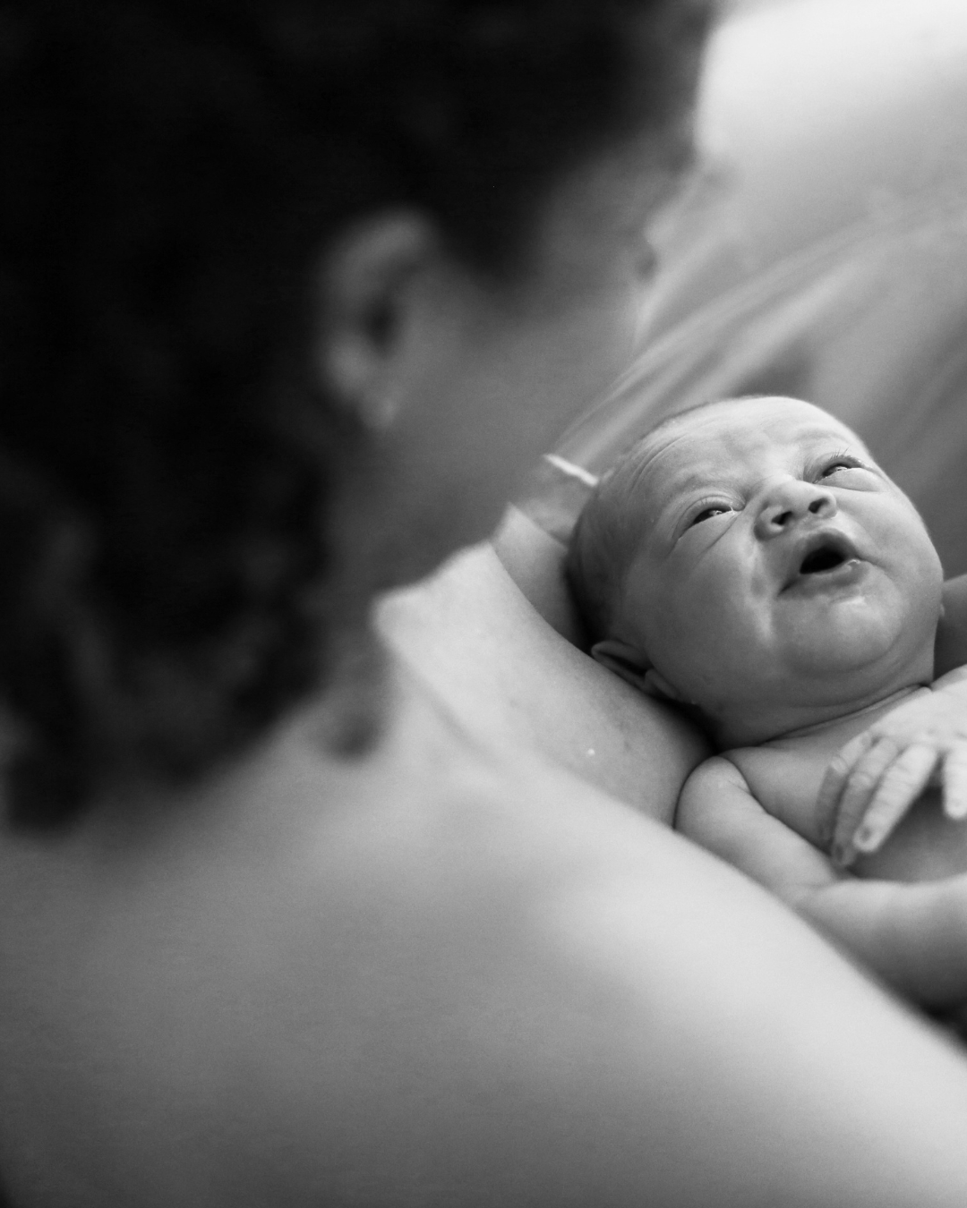 A black and white photo of a newborn baby lying on an adult's chest, gazing upwards with open eyes, while an adult's face is partially visible in the foreground, slightly out of focus.