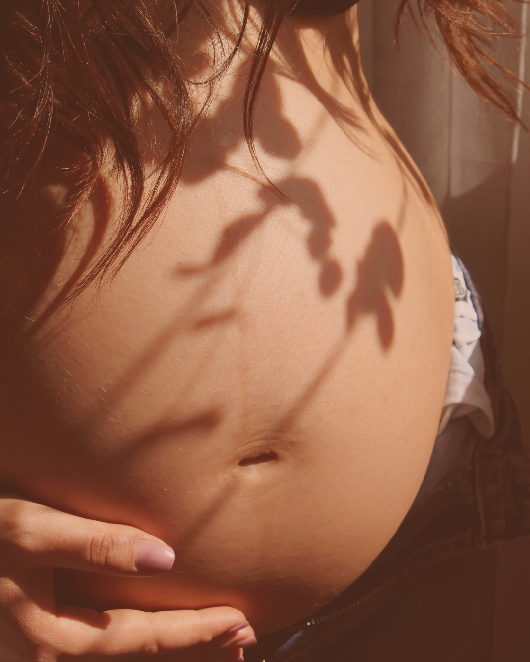 Close-up of a pregnant woman's belly with shadow of a leaf on it, her hand touching her abdomen, sunlight highlighting her skin.