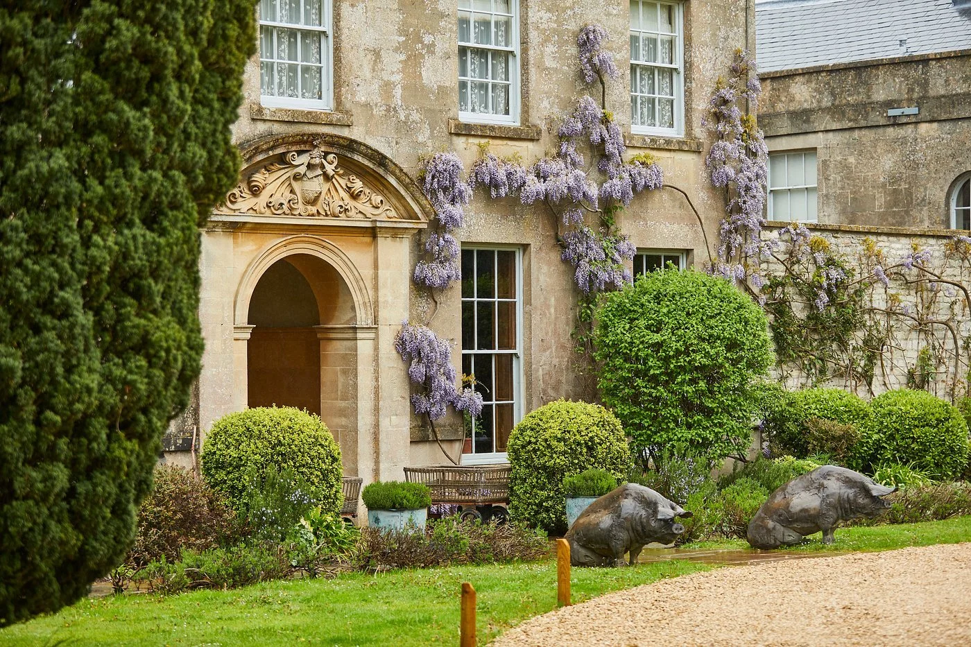 Garden with blooming purple wisteria on a stone house, decorative sculpted entrance, trimmed bushes, and pig sculptures on the lawn.