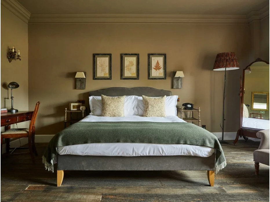 A bedroom with a bed centered against a beige wall, three framed botanical prints above it, two wall-mounted lamps on either side, a wooden desk with a chair to the left, a tall floor lamp with a red shade on the right, a standing mirror tilted against the wall, and a gray armchair partially visible in the corner.
