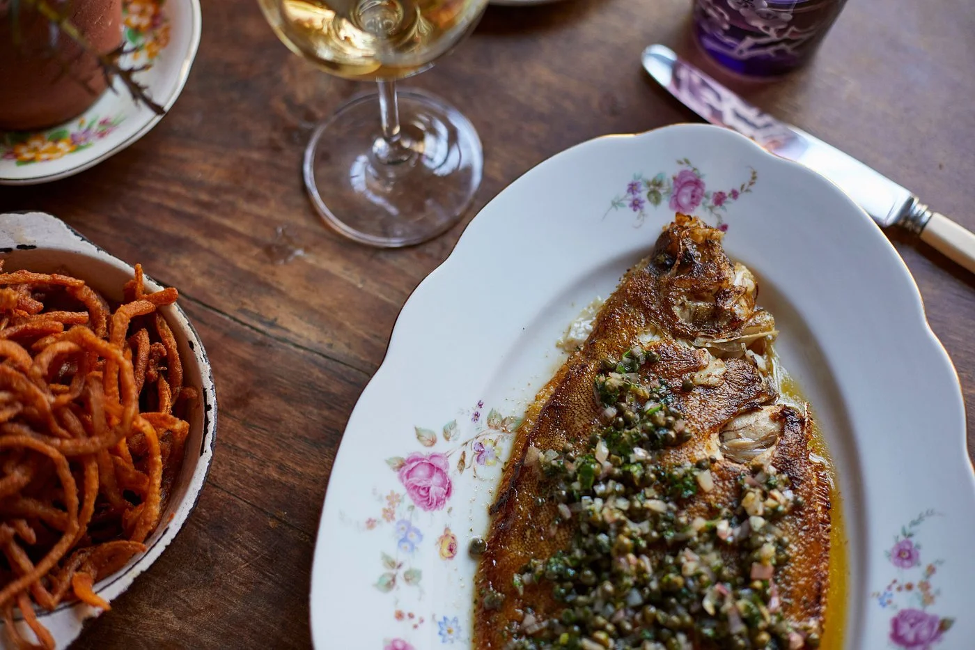 A seafood dish with a whole fish topped with chopped herbs and garlic, served on a floral porcelain plate on a wooden table, with a glass of white wine, a bowl of spiral pasta, and utensils nearby.