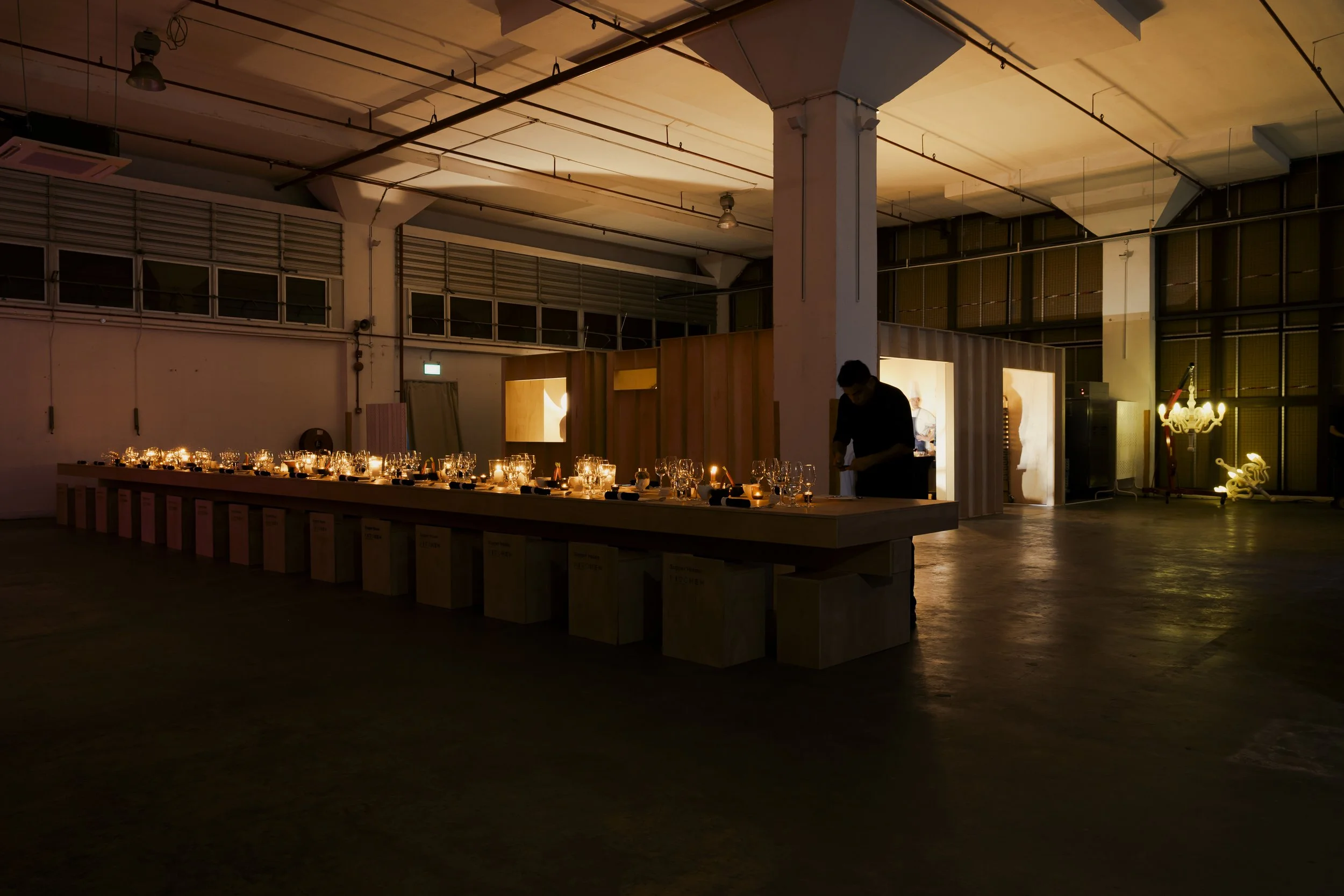 Dimly lit event space with a long table set with candles, glasses, and tableware, and a person standing nearby, with some illuminated art or decorations in the background.