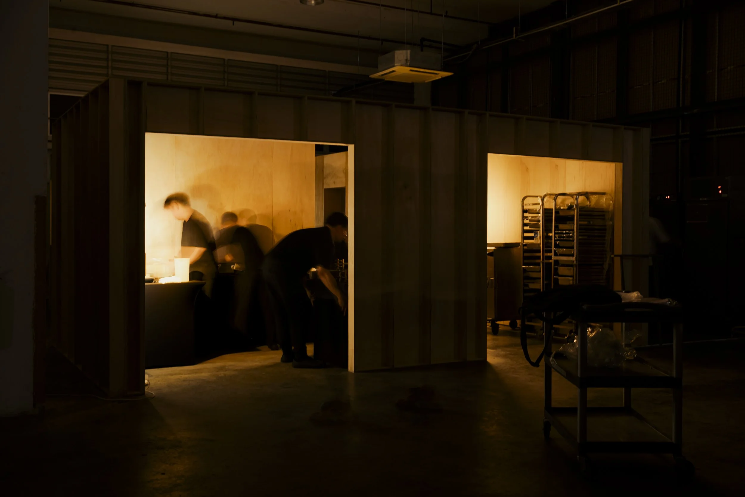 Dimly lit room with a wooden partition, several people working or standing around, carts and stacked trays, possibly a kitchen or preparation area.