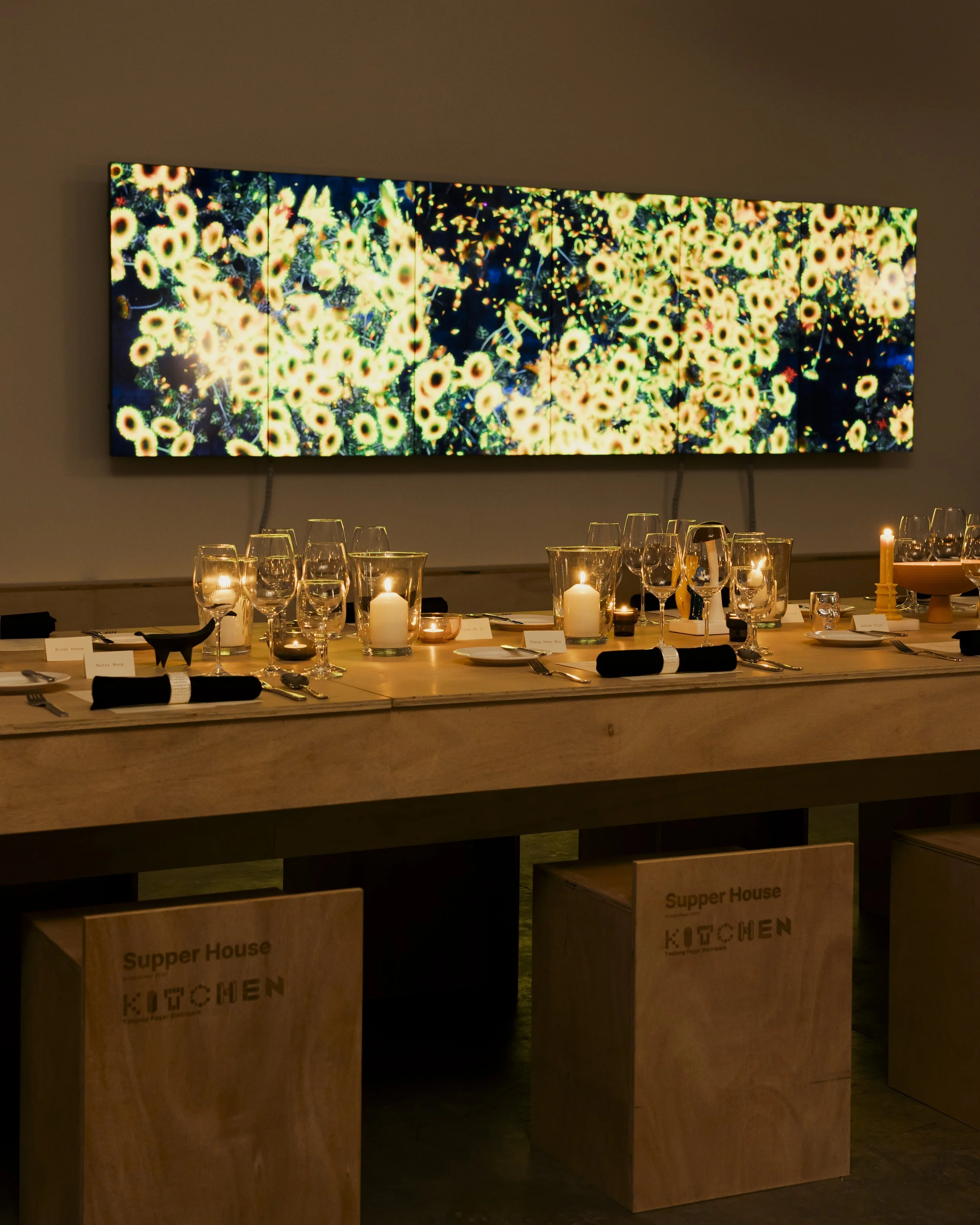 A wall-mounted display showing an abstract image of many yellow and black circular shapes, resembling a field of flowers, in a dimly lit room above a table set with candles and glasses.