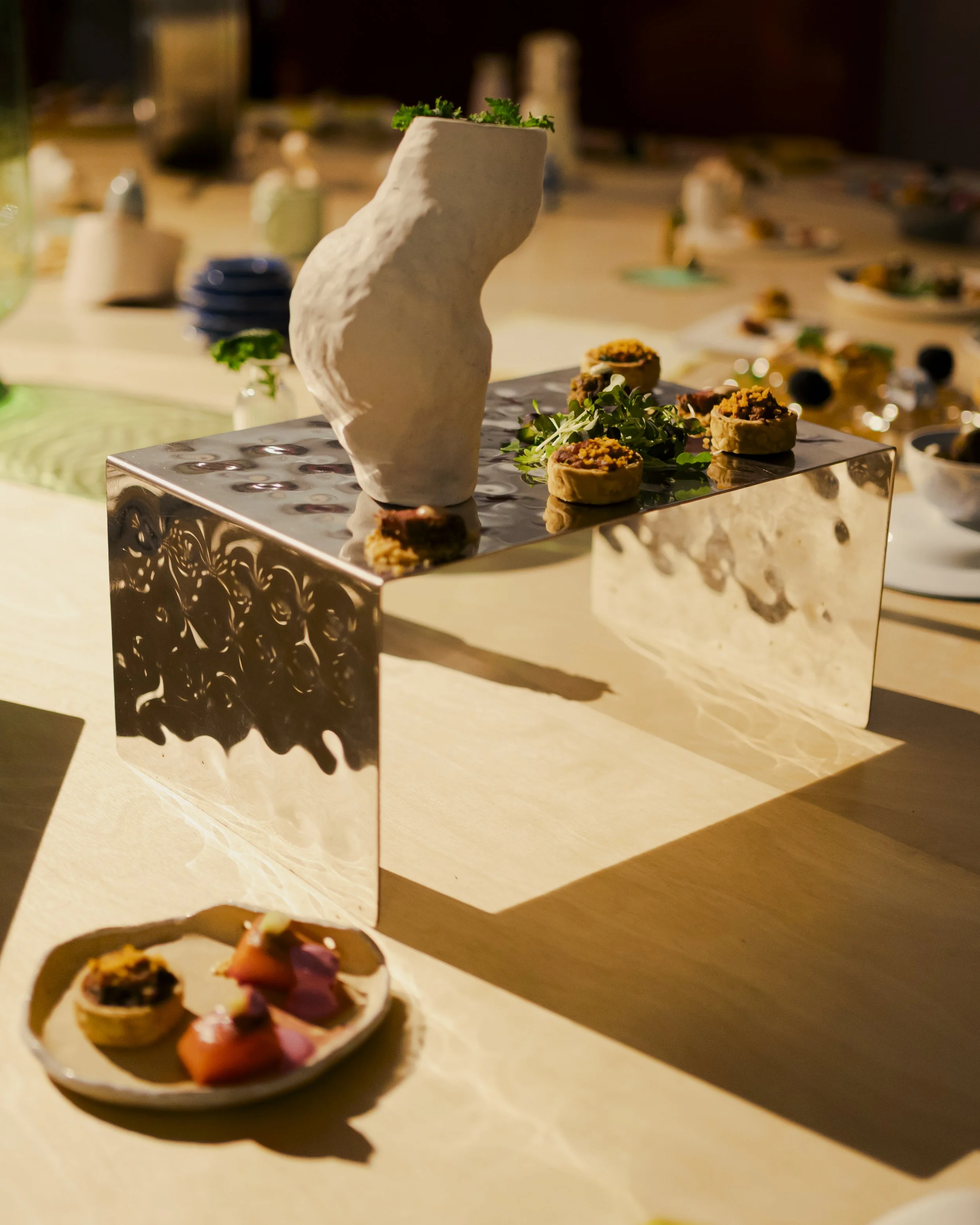 Close-up of a modern decorative sculpture on a table with small food dishes in the background. The sculpture has an abstract, textured, cream-colored form with a small plant at the top. The table has a shiny, reflective surface with intricate pattern