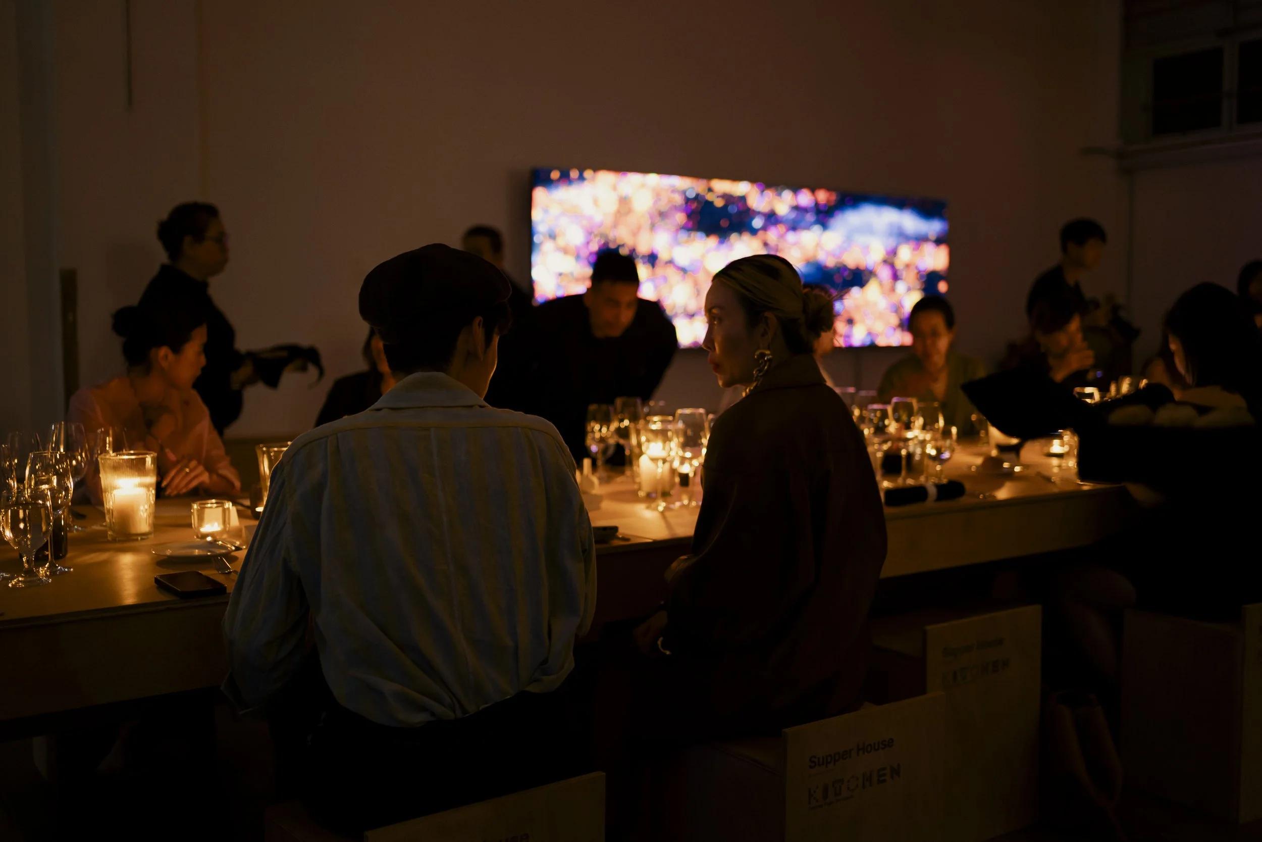 People sitting at a dinner table with lit candles in a dimly lit room, with a large, colorful, abstract digital display on the wall behind them.