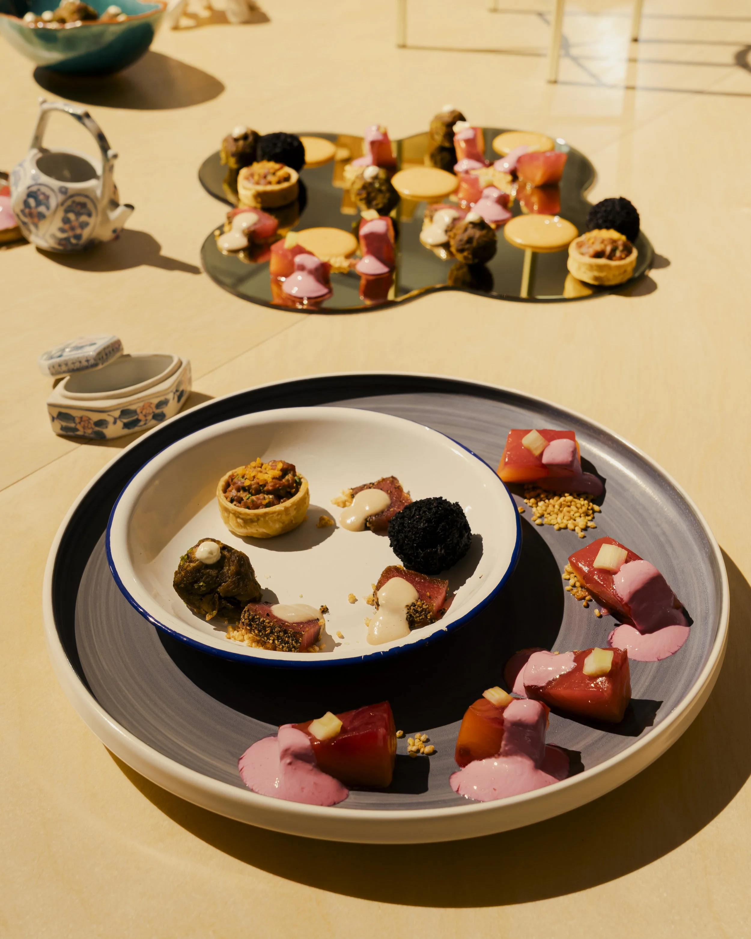 Assorted gourmet desserts on a white and black plate, including small tartlets, chocolate truffles, and pink and yellow confections, with additional desserts and a ceramic teapot in the background.