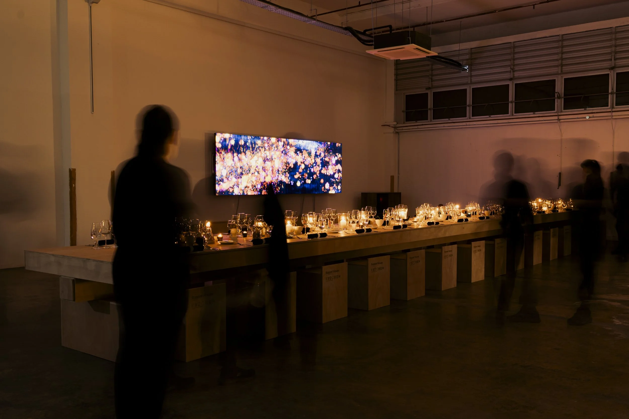 A dimly lit event space with a long dining table set with wine glasses and candles. A flat-screen TV displaying colorful abstract visuals is mounted on the wall, and there are silhouettes of people standing and walking around.