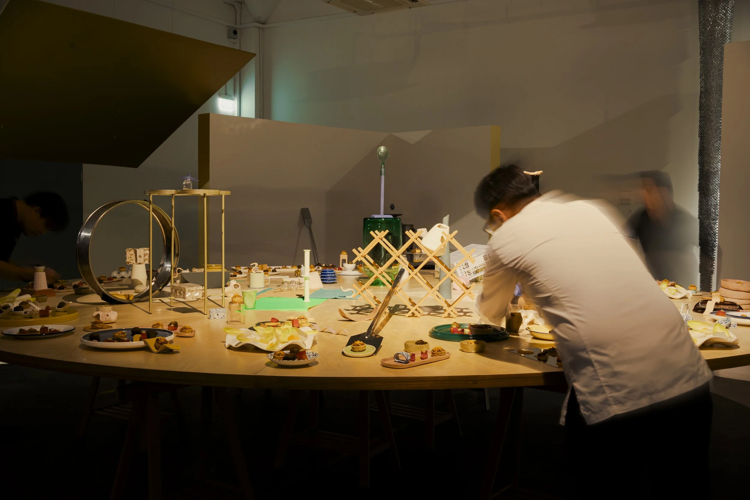 A person wearing glasses and a white shirt is leaning over a table filled with various small decorative objects, dishes, and desserts. The table also has some creative sculptures and art pieces, with a blurred background of an indoor space.