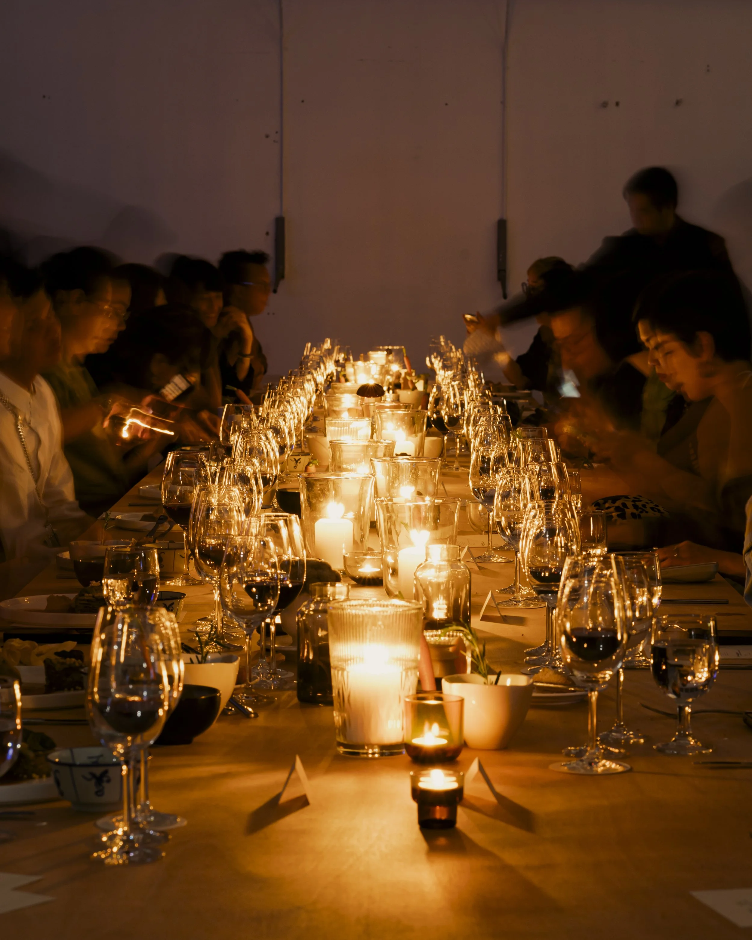 A long dinner table with lit candles and wine glasses, surrounded by people in a dimly lit setting.