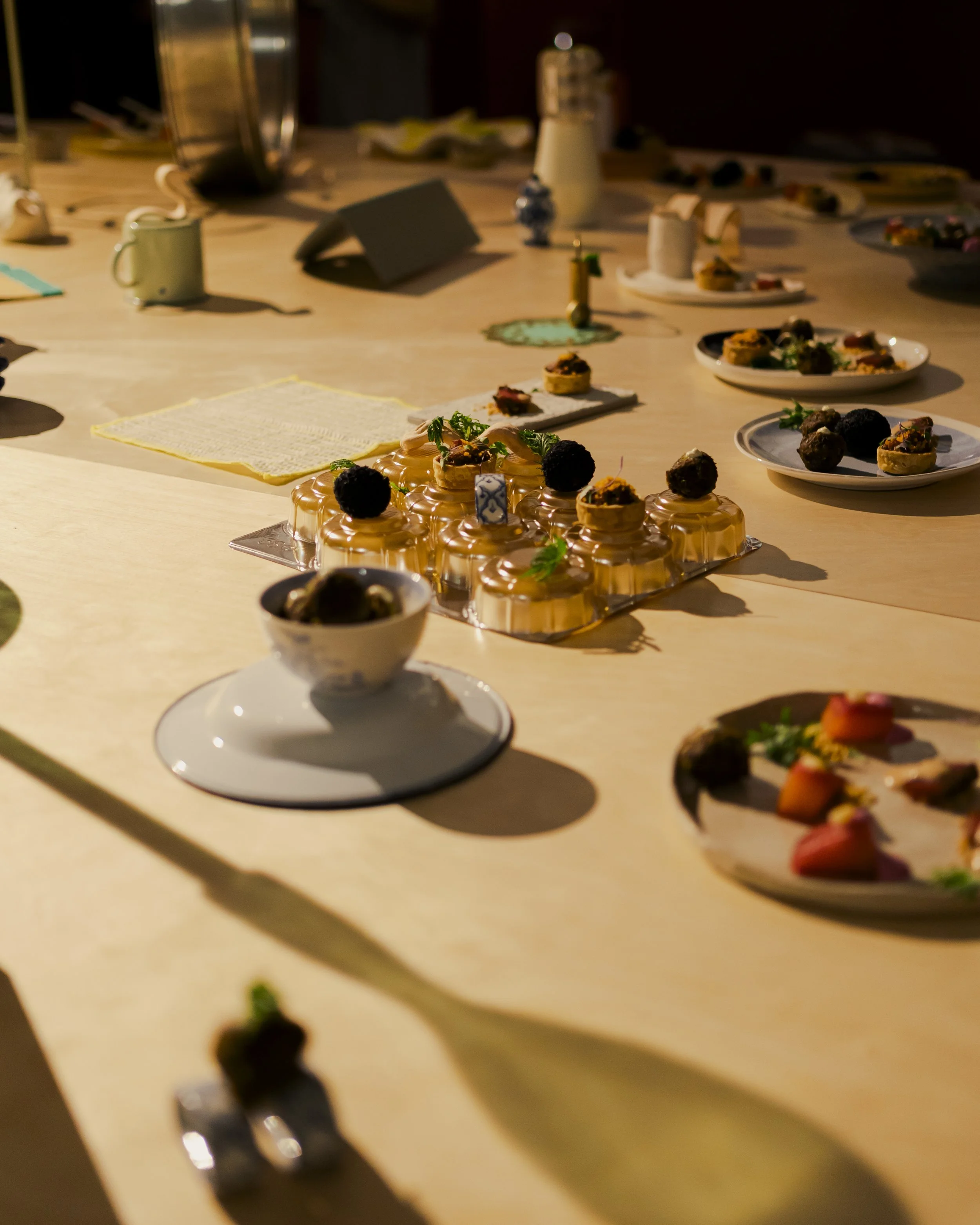 A variety of desserts on a table, including mini cakes topped with berries and garnishes, and plates with assorted small pastries and treats, under warm lighting.