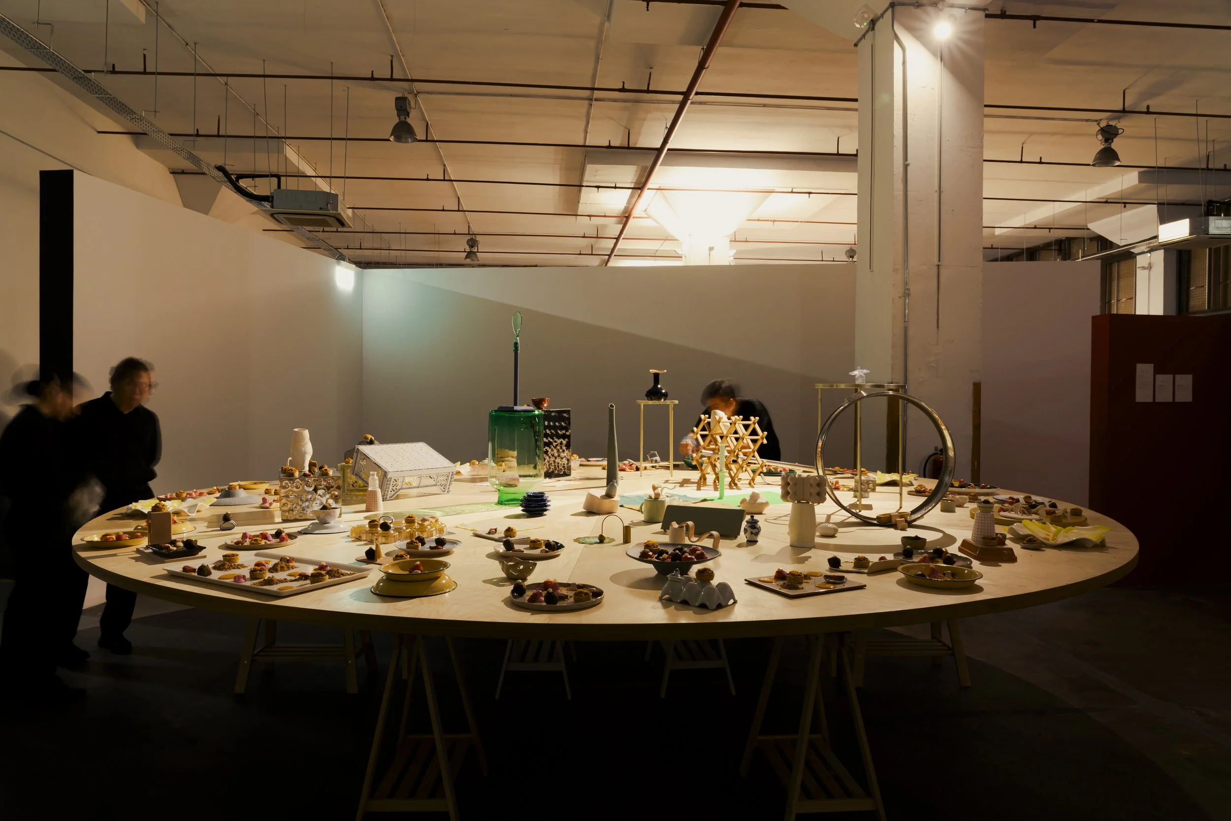 Large round table displaying various ceramic and decorative objects, with people examining them in a spacious industrial-style gallery with high ceilings and exposed pipes.