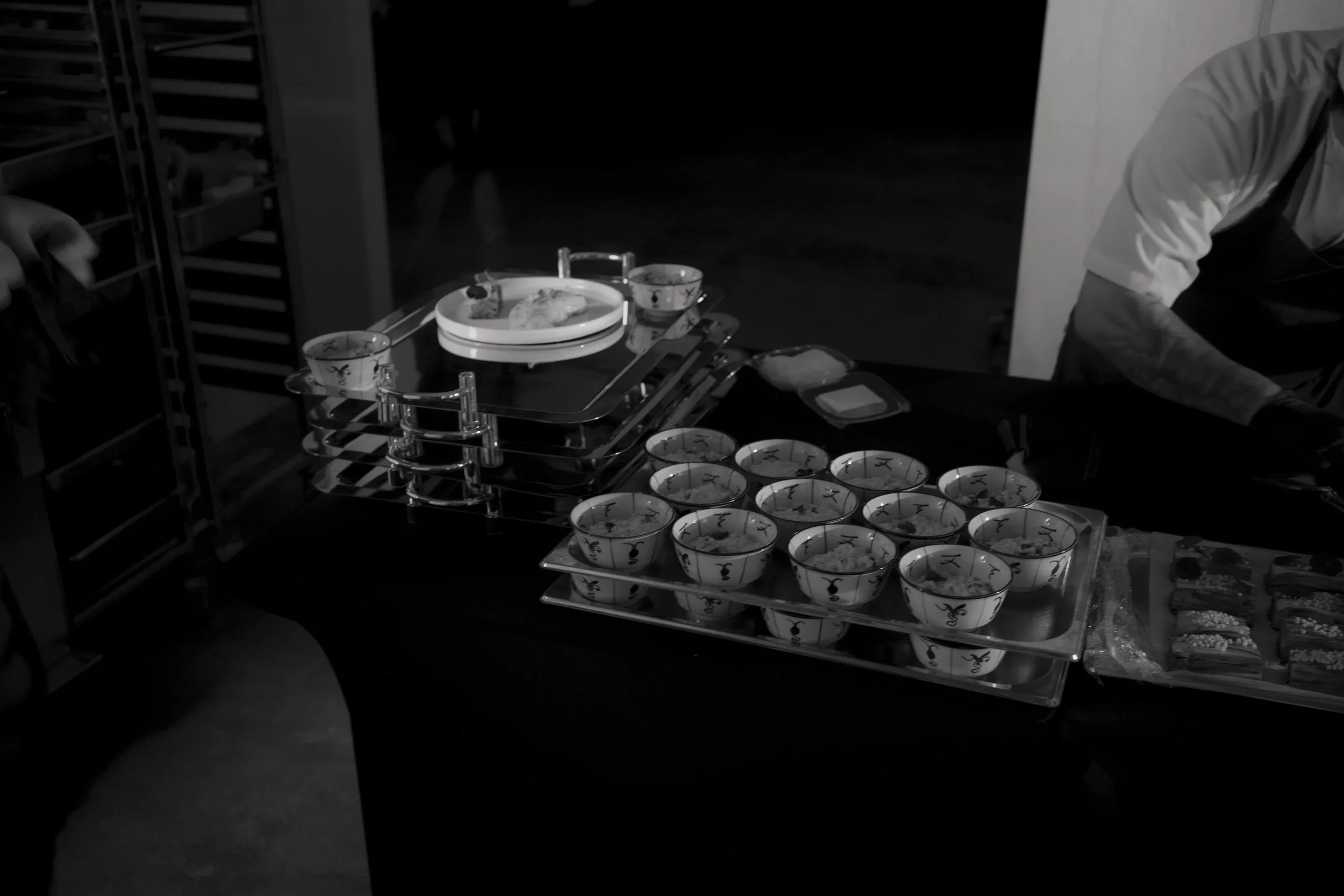 A catering table with small patterned bowls filled with food, several cups, and a tray with a piece of bread or pastry, in a dimly lit setting.