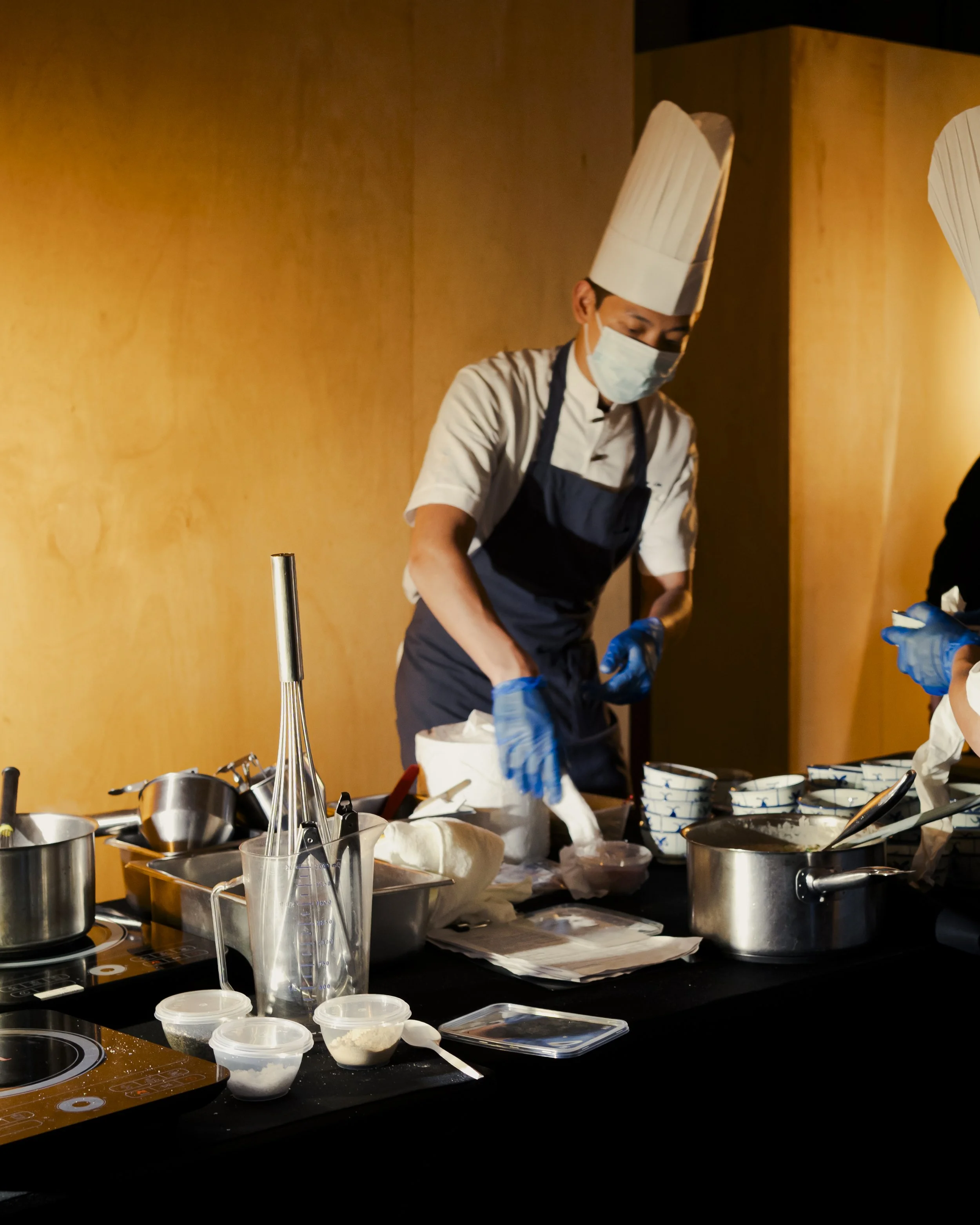 A chef wearing a tall white hat, face mask, and apron is working at a kitchen counter with various utensils, bowls, and ingredients around him.