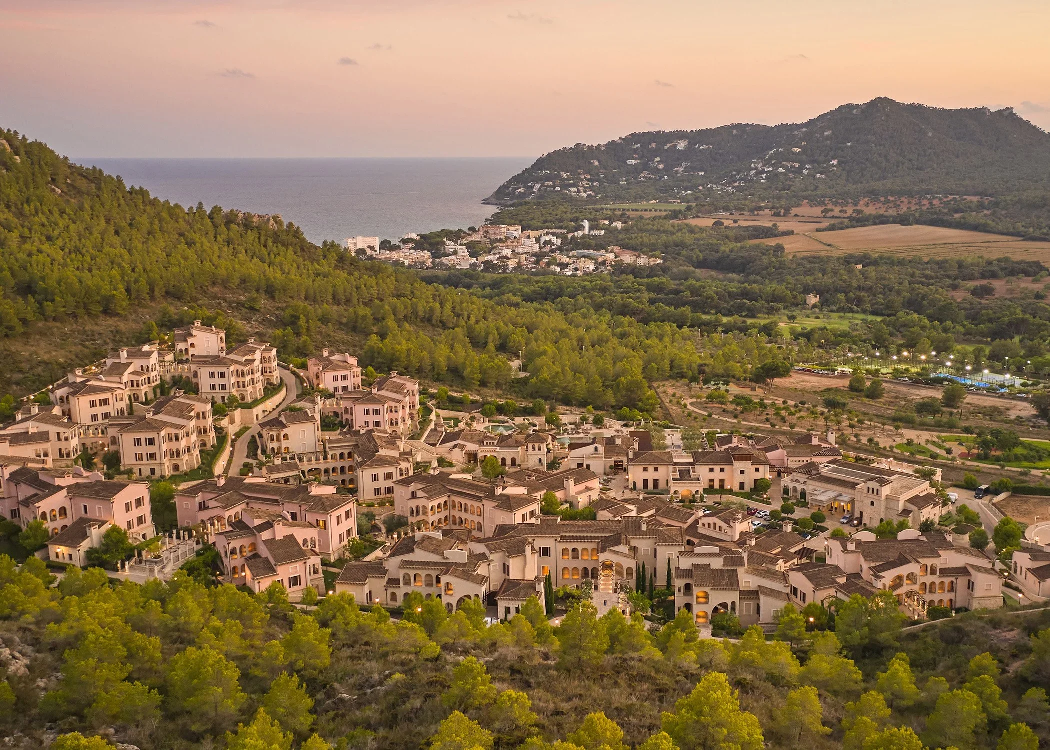 Aerial view of a coastal town with rows of white residential buildings, surrounded by lush green trees, hills, and a large body of water in the background at sunset.