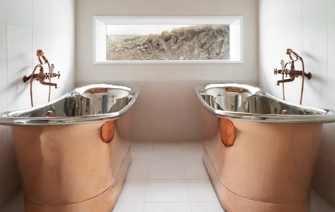 Two copper bathtubs with copper fixtures in a bathroom with a large horizontal window showing a rocky landscape outside.