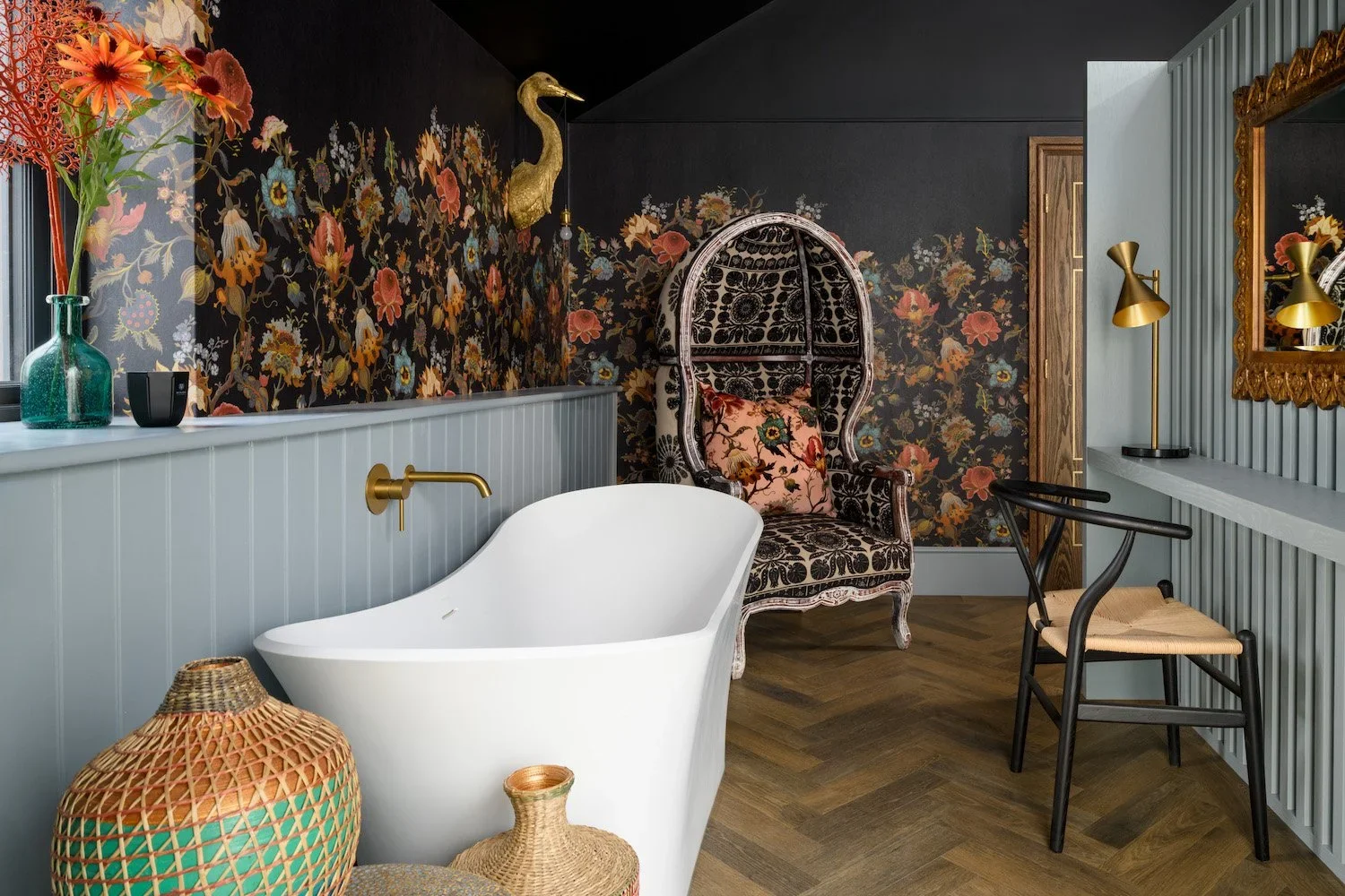 Bathroom with floral wallpaper, a white freestanding bathtub, and black-and-white patterned armchair with a floral cushion, decorated in a vintage style.