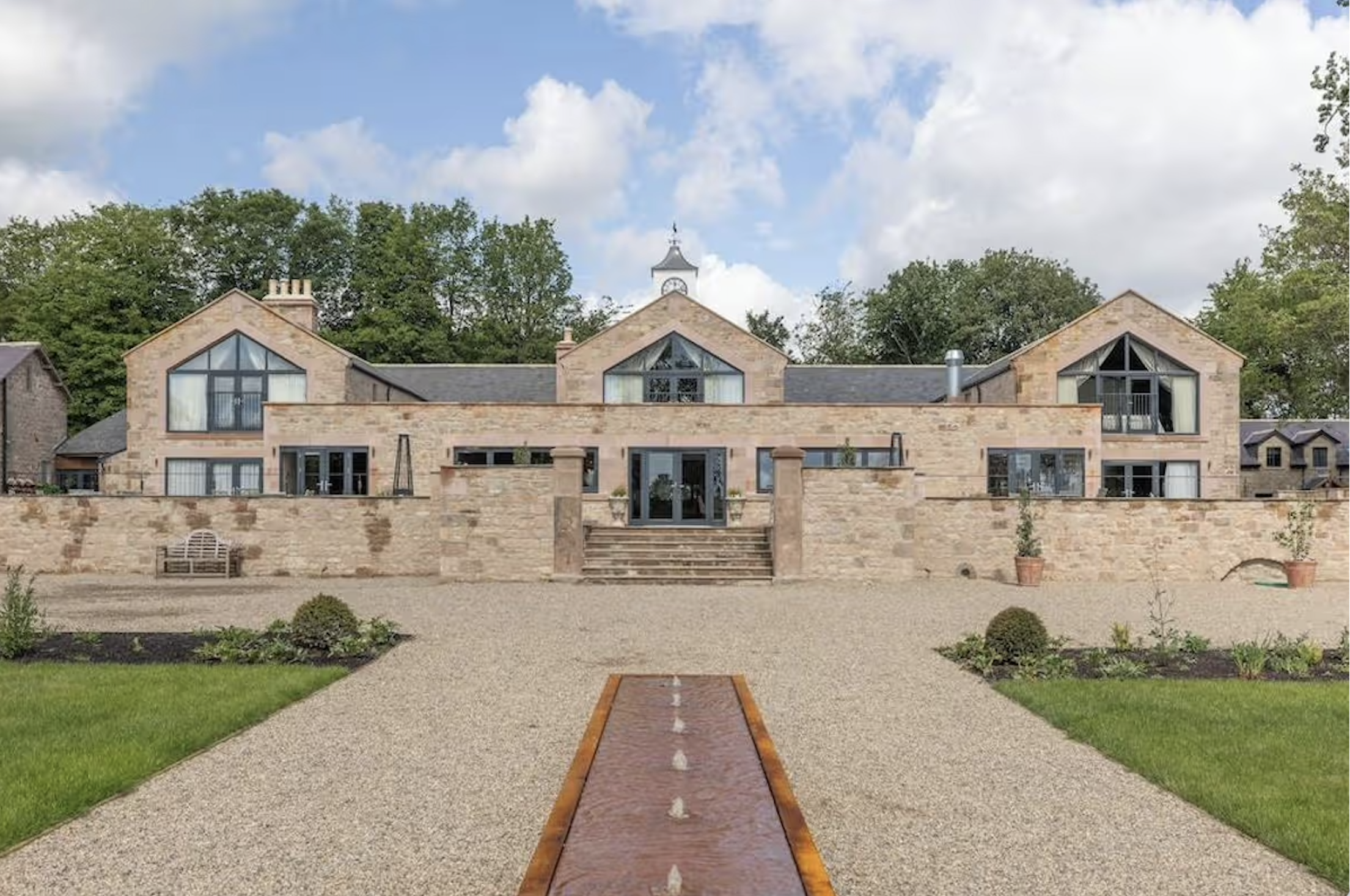 Stone house with large glass windows and a clock tower surrounded by trees, with a gravel driveway and steps leading up to the entrance.