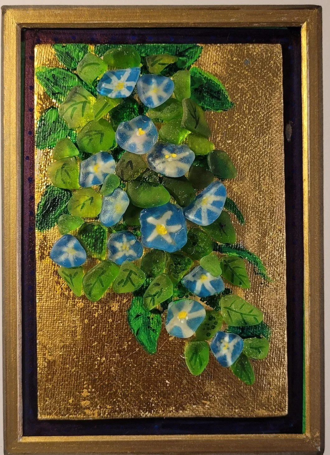 A framed artwork featuring small glass flowers and leaves set against a gold textured background.