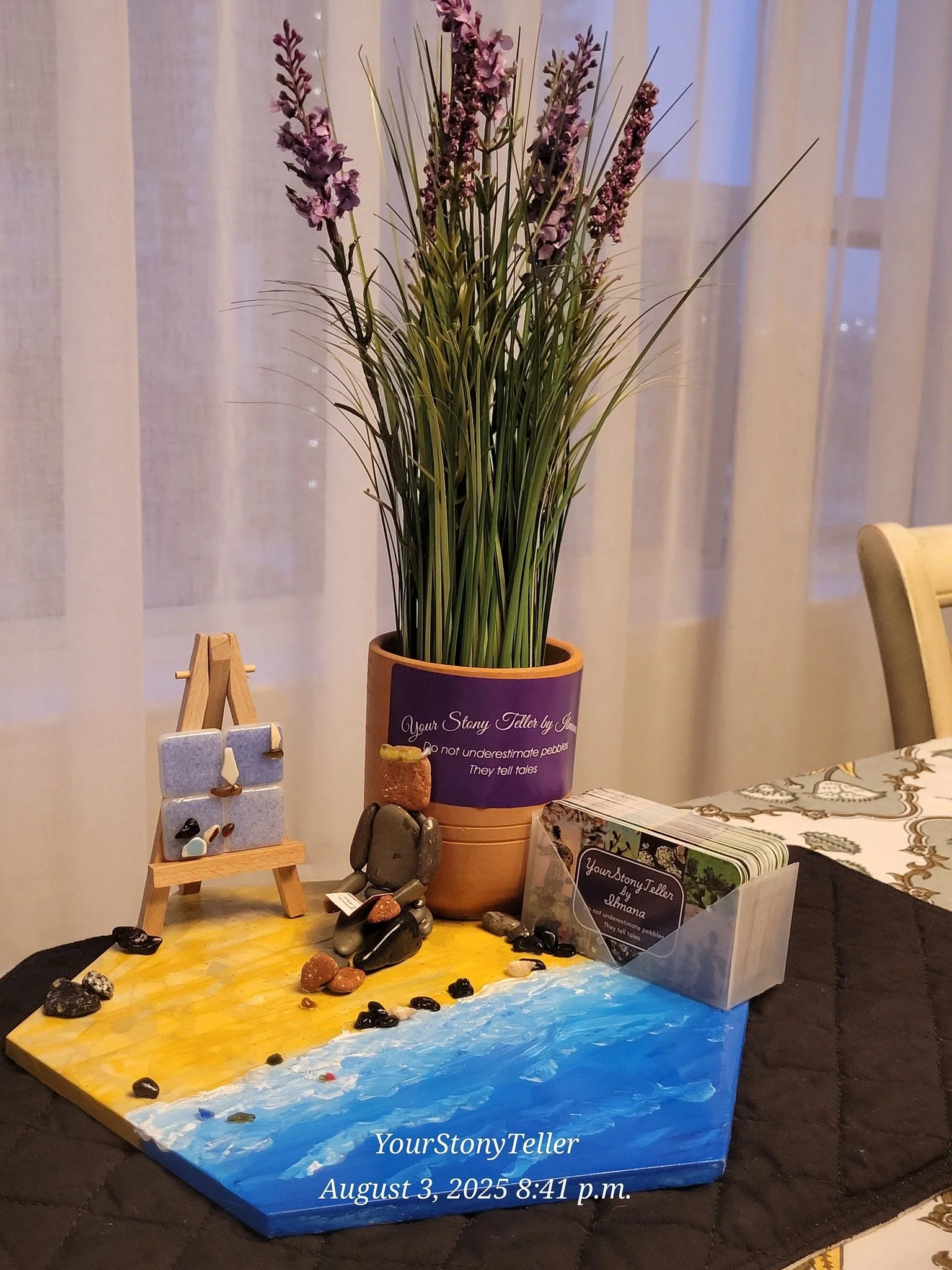 Decorative tabletop scene with a potted lavender plant, rocks, a small easel with painted stones, and a deck of cards in a holder. The table has a painted beach scene with a timer showing the date August 3, 2025, and the time 8:41 p.m.